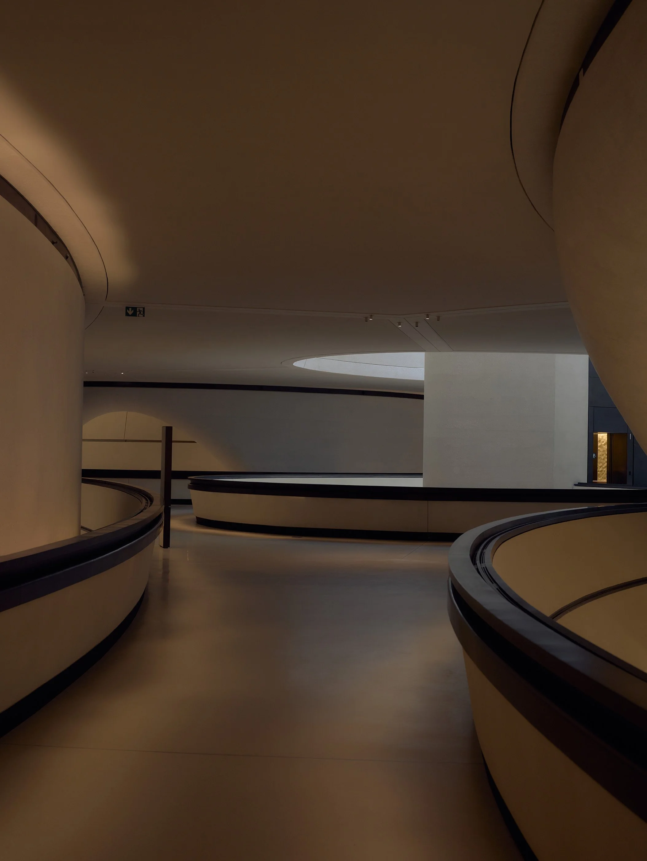 A modern interior with curved, beige walls and a winding, dark railings along a staircase or walkway. The lighting is subdued, creating a calm atmosphere, with a small rectangular window or opening visible on the right side.