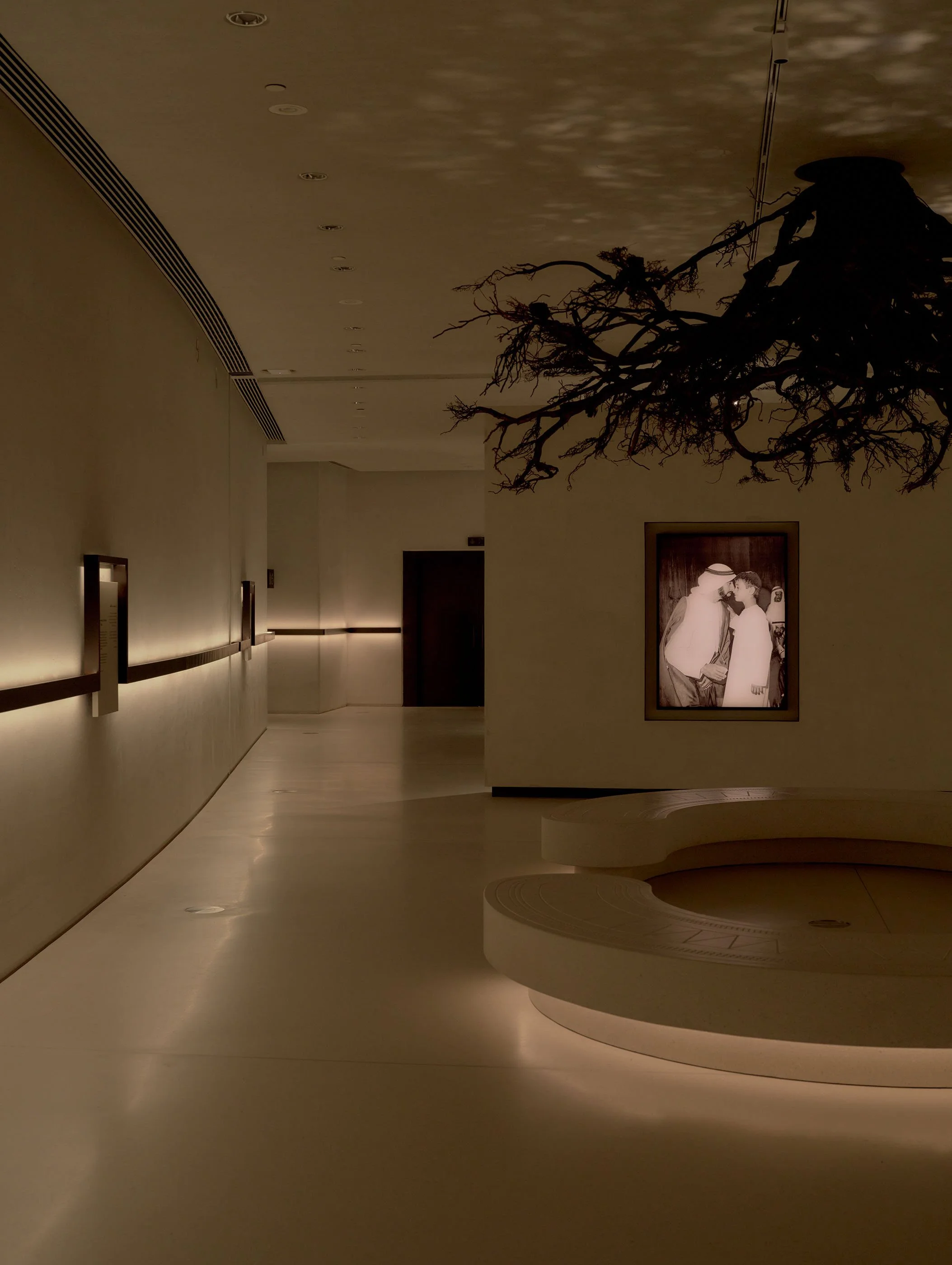 A modern, minimalist hallway with beige walls and a curved, built-in bench. The ceiling has small, recessed lights. There is a large framed photograph of two men, possibly in traditional Middle Eastern attire, on the wall to the right. A bare tree br