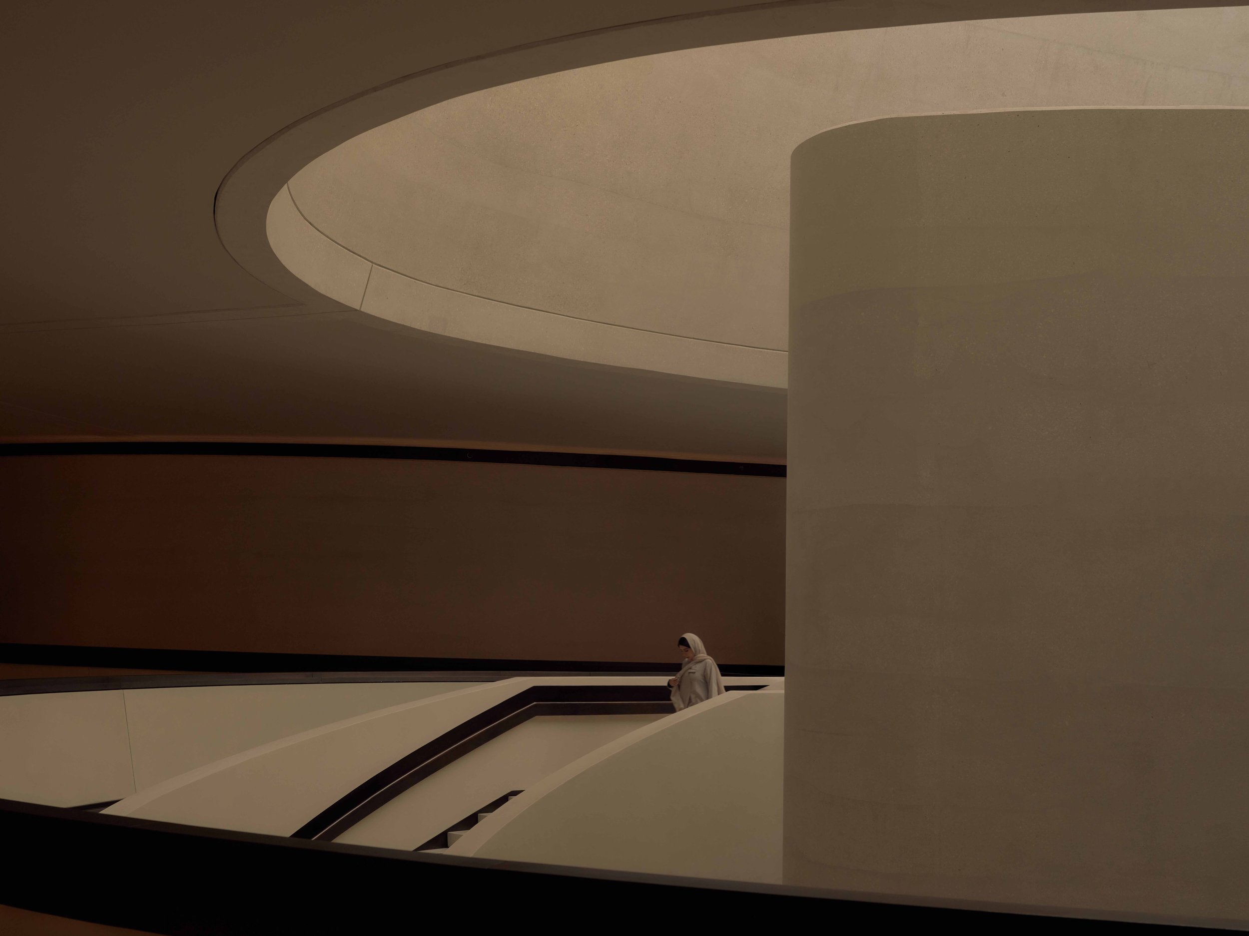 Modern interior architecture with large curved concrete walls and a staircase. A person dressed in white is walking up the stairs.