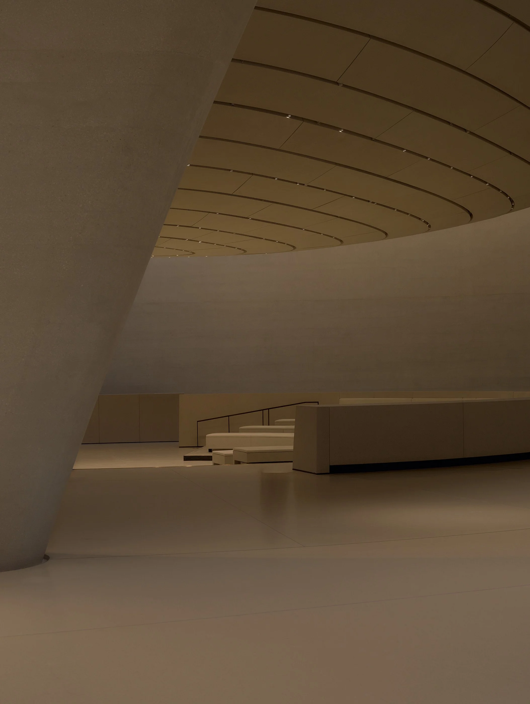 Interior view of a modern building with curved ceiling panels, minimalist white benches, and soft lighting.
