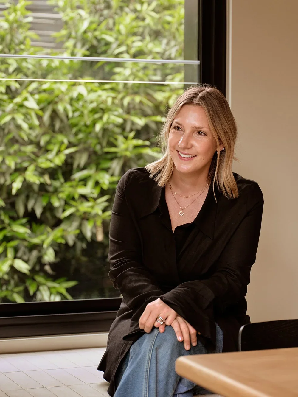 A woman with shoulder-length blonde hair sitting by a large window with greenery outside. She is smiling and wearing a black shirt and jeans, accessorized with layered necklaces and rings.