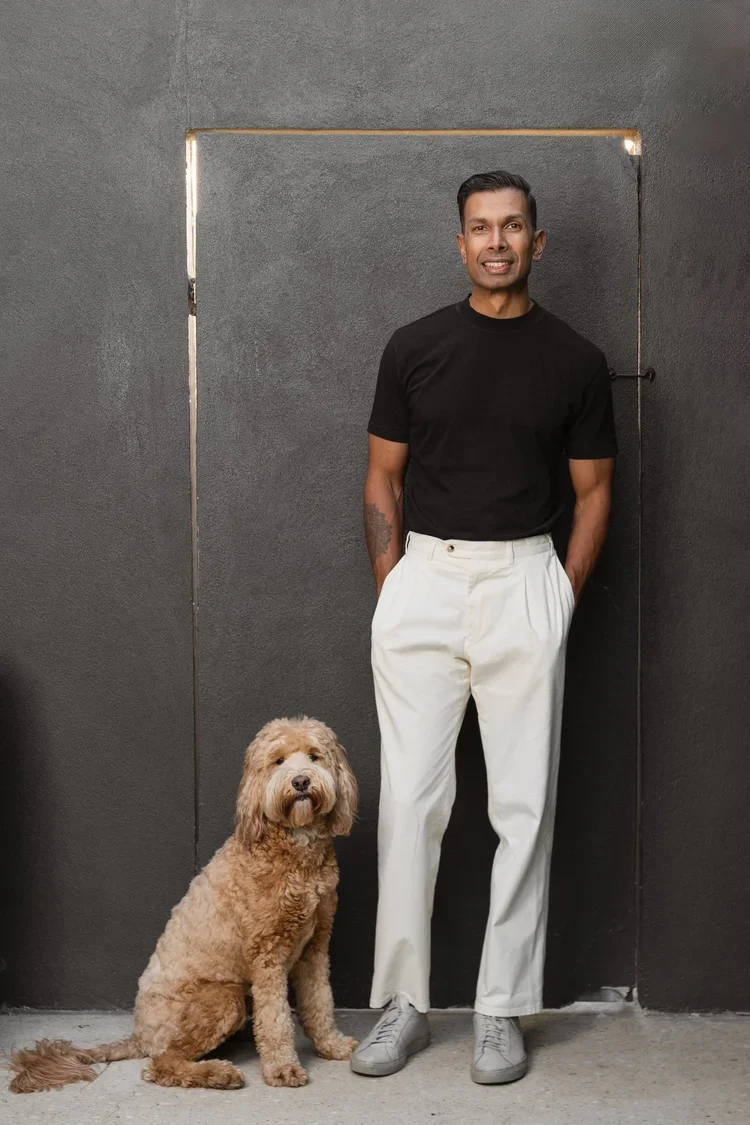 A man standing with his hands in his pockets next to a curly-haired dog in front of a gray wall.