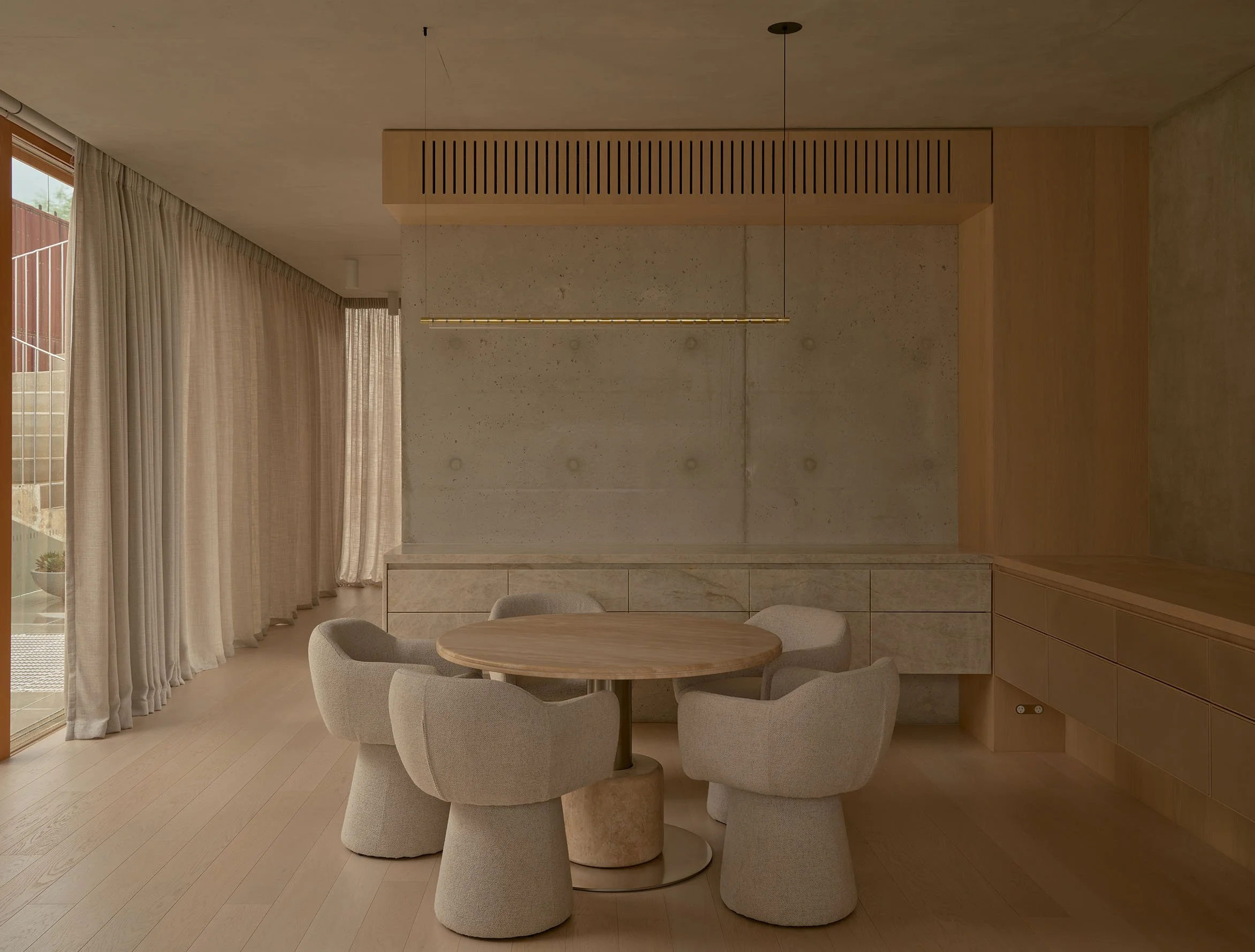 Modern dining area with a round wooden table and six beige upholstered chairs; large window with beige curtains; concrete wall with a wooden beam and built-in cabinetry.