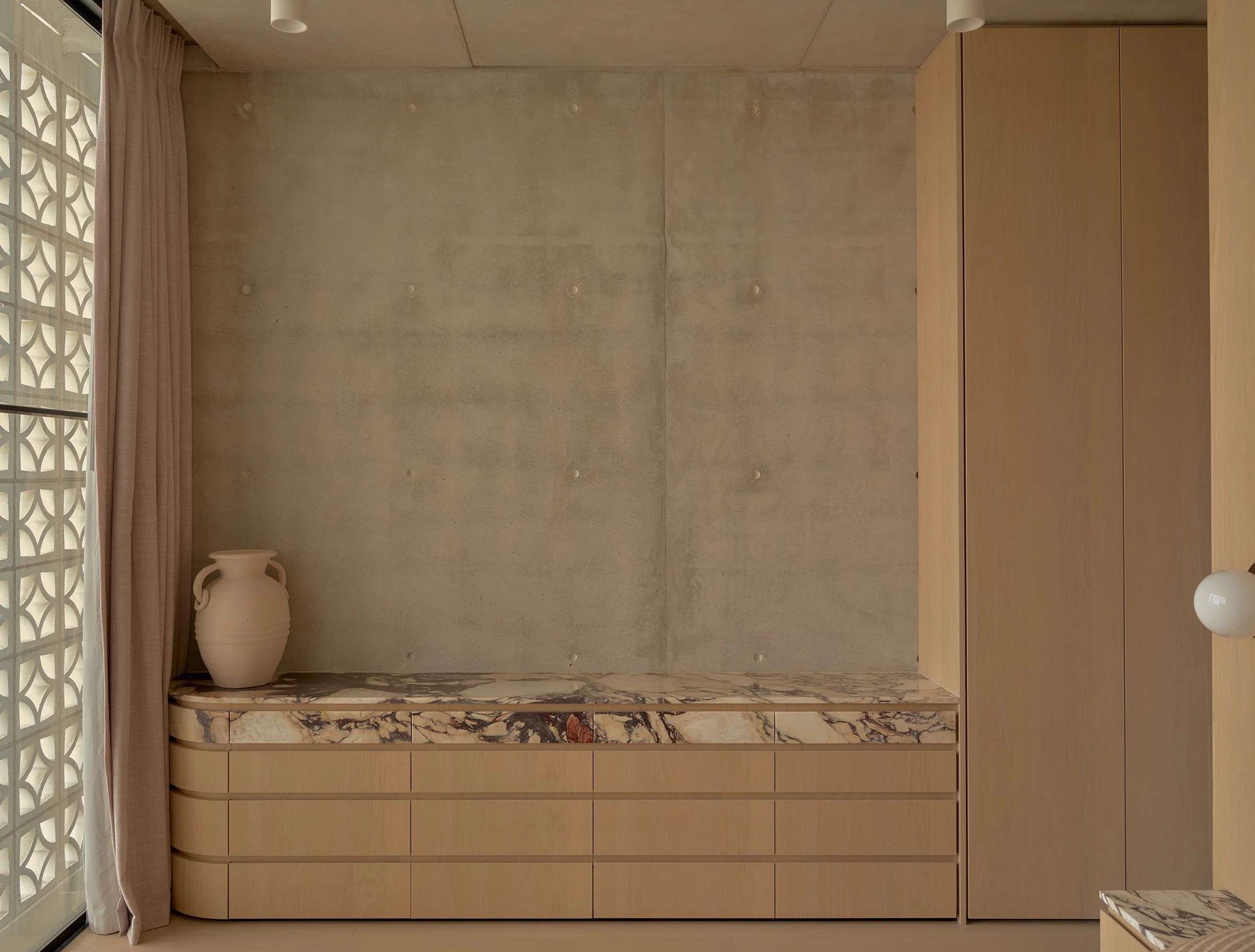 Interior of a modern room featuring a wooden cabinet, a marble countertop with a white vase, pink curtains, a decorative window, and a light-colored wall with round button details.