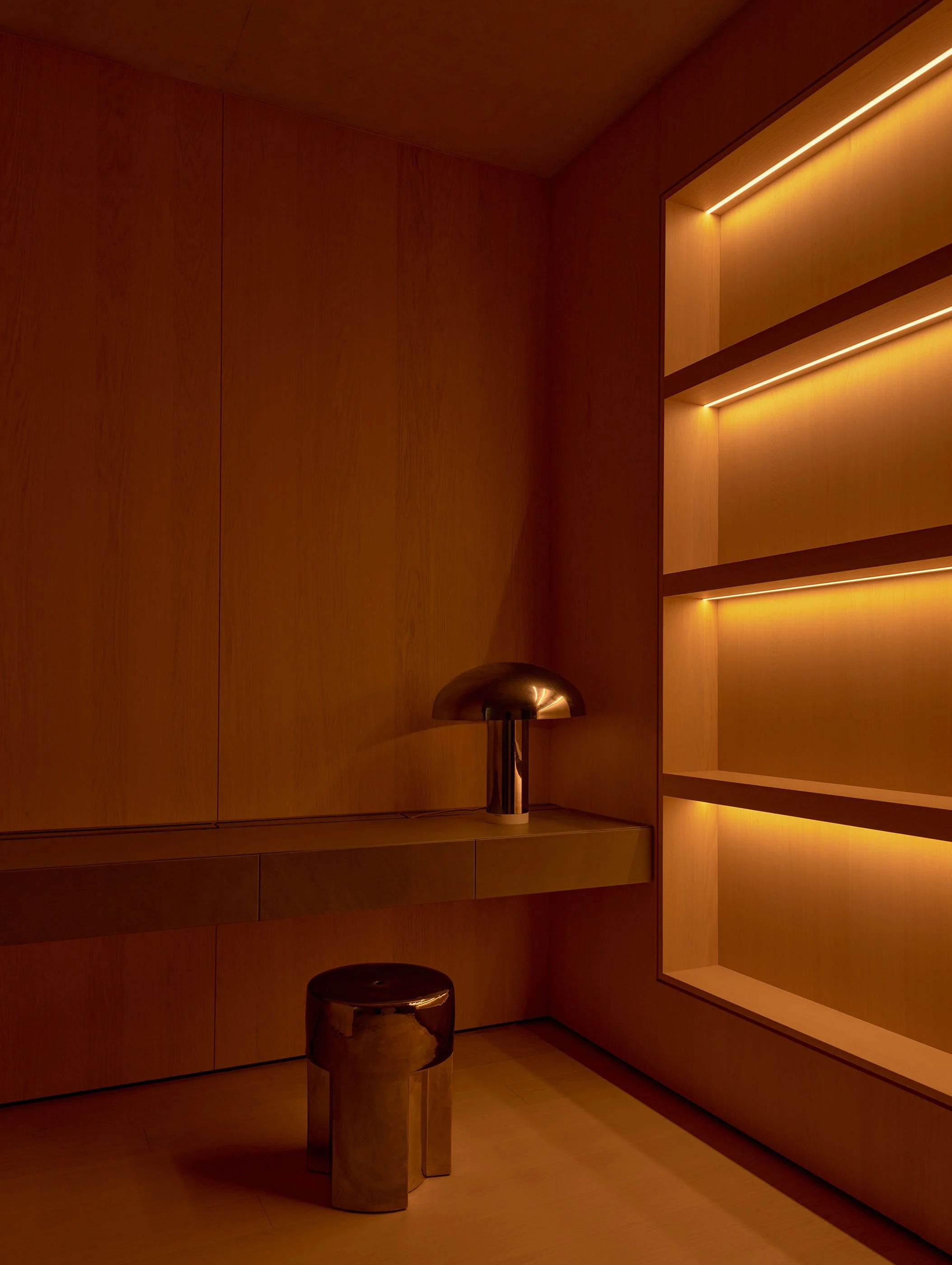 A wooden interior with built-in shelves illuminated by warm lighting. There is a metallic table lamp on a wooden surface and a shiny, rounded stool below.