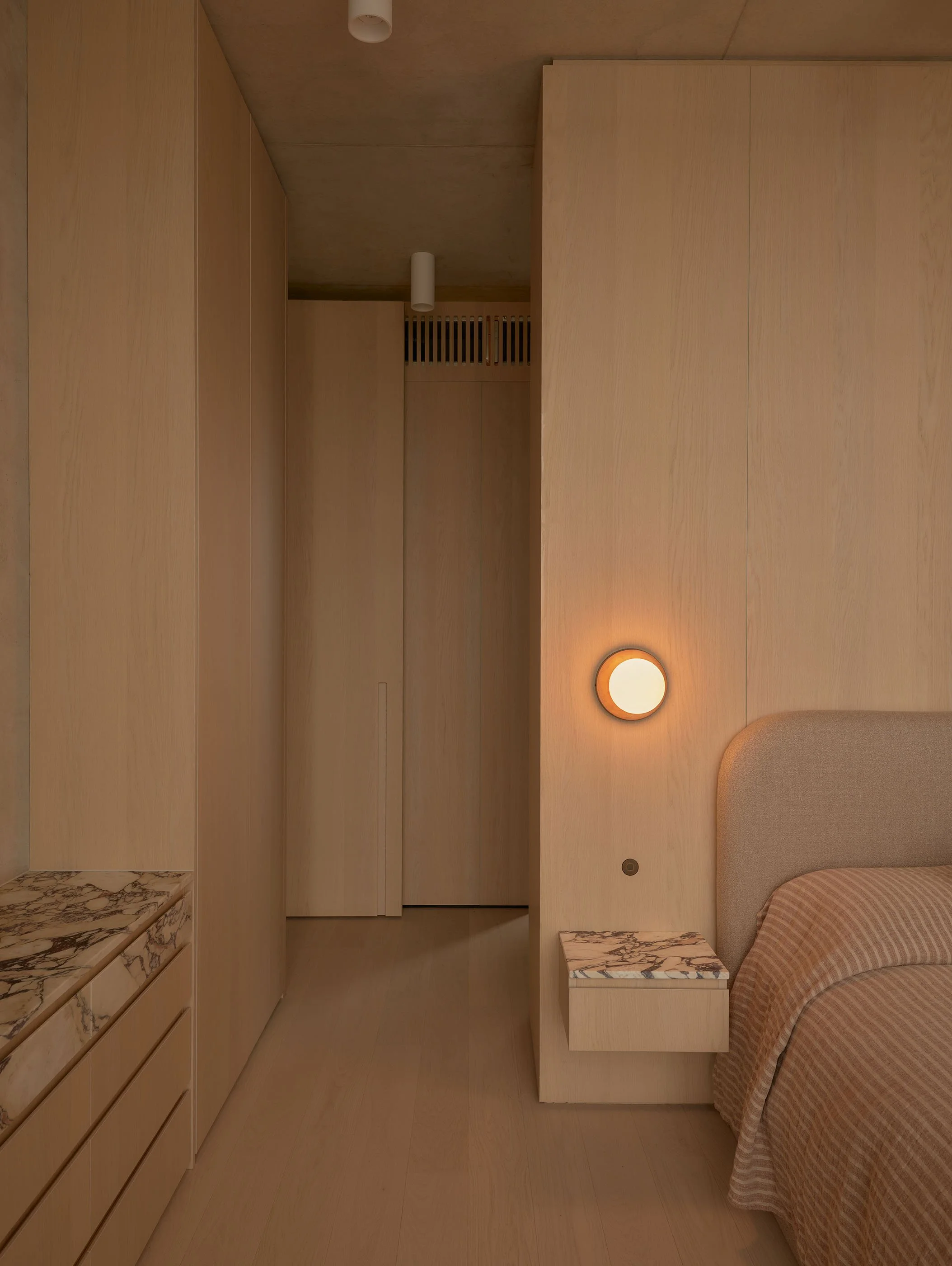 A bedroom corner with a bed, a built-in wardrobe with wooden panels, a marble top cabinet, and a ceiling light fixture.
