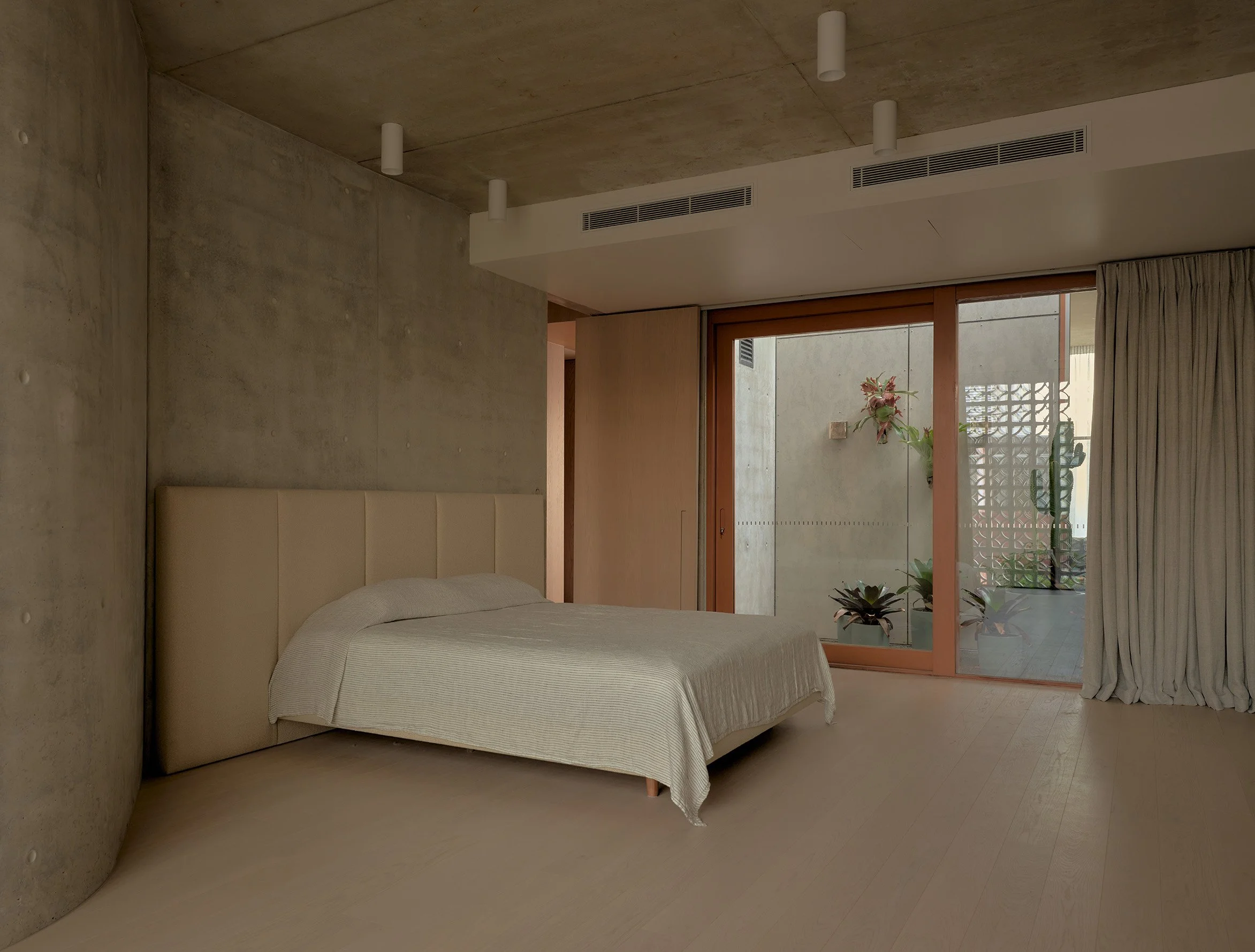 A bedroom with concrete walls and ceiling, a bed with a light-colored bedspread, a large window with wooden framing, beige curtains, potted plants near the window, and ceiling-mounted lights.