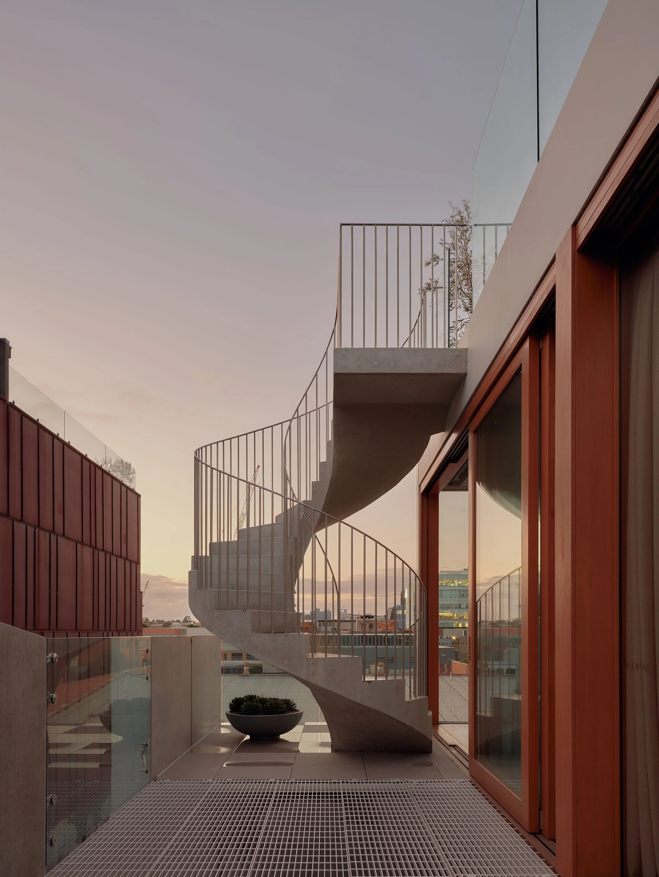 A modern building balcony with a spiral concrete staircase, glass railing, and wooden window frames during sunset.