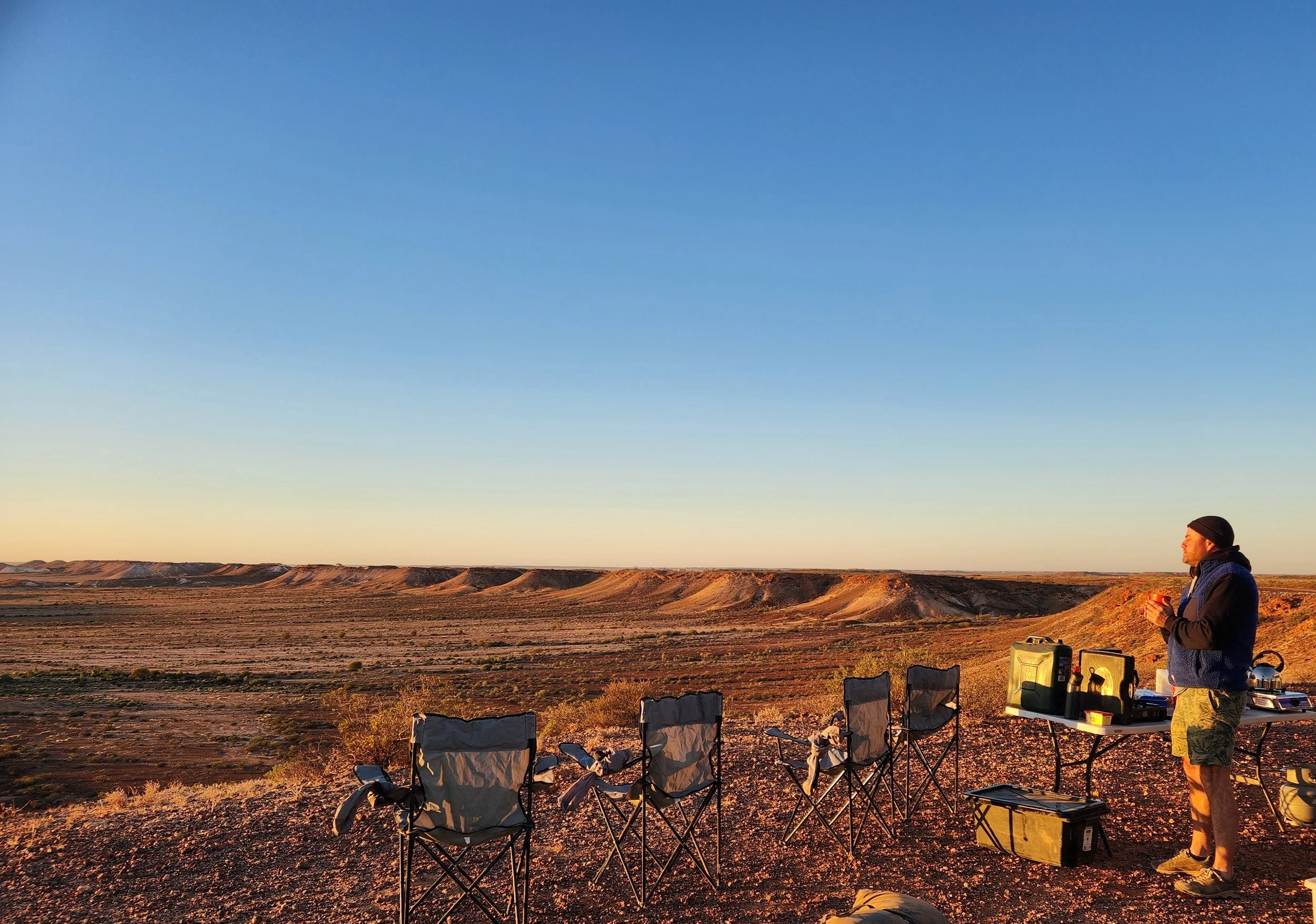 A person standing near camping chairs and gear in a desert landscape during sunset, with distant hills and a clear sky.