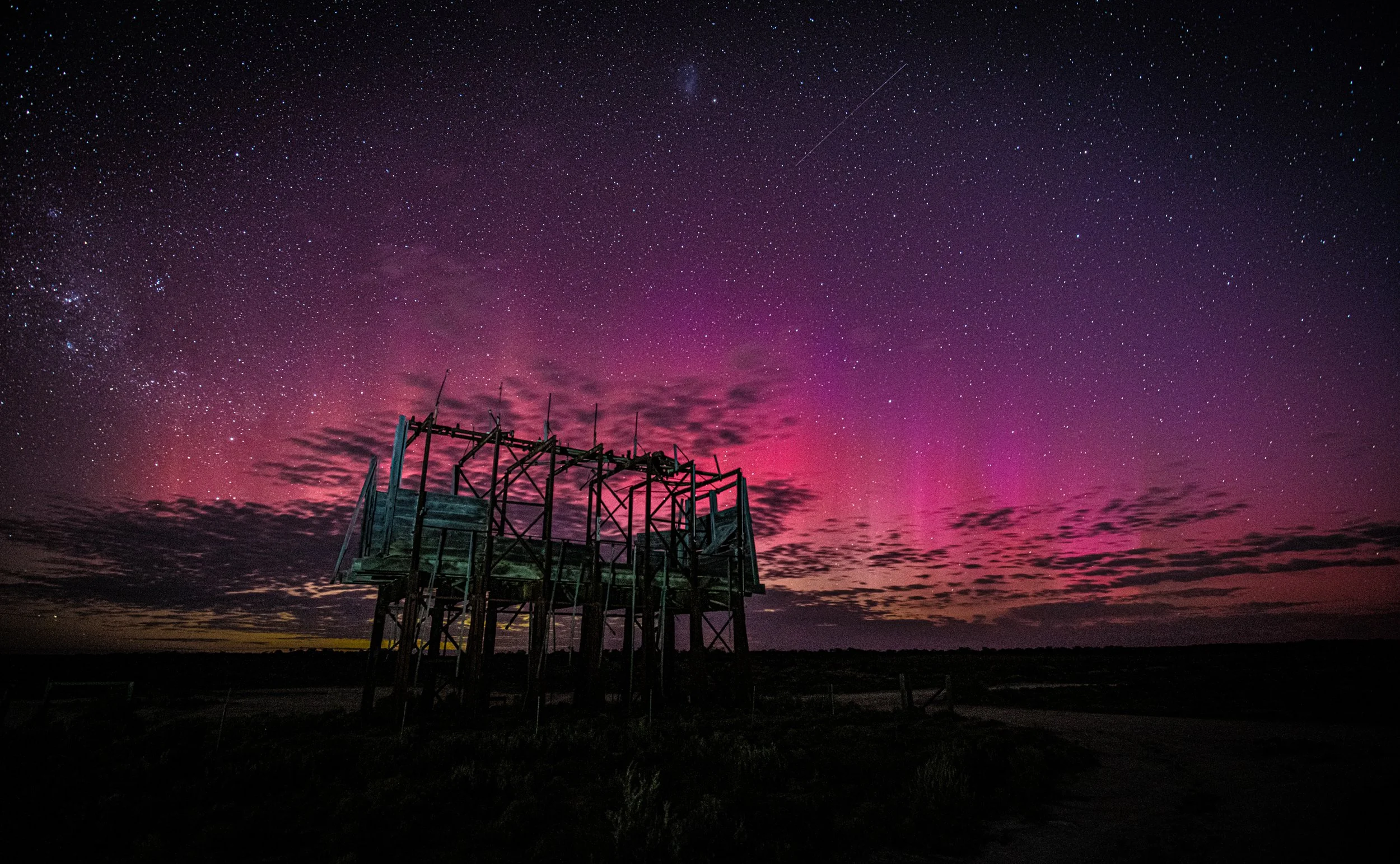 An abandoned wooden observation tower on a flat landscape against a vibrant purple and pink night sky filled with stars, clouds, and a shooting star.