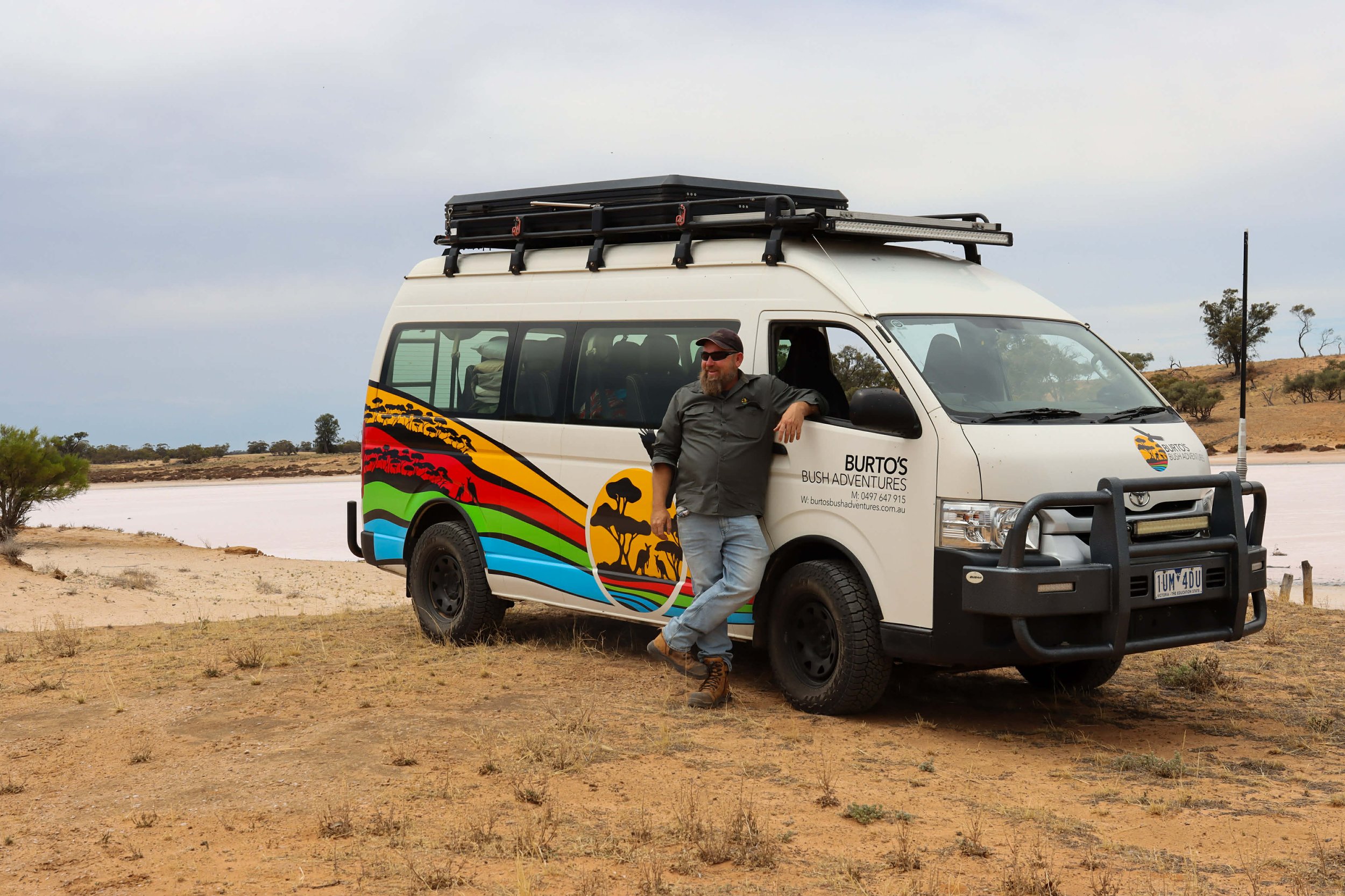 A man with a beard, sunglasses, gray shirt, and jeans leaning against a colorful adventure van with a scenic landscape, lake, and cloudy sky in the background.