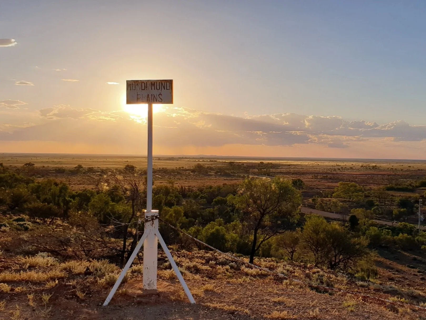 A landscape scene with a sunset over a semi-arid plain, featuring a sign that reads 'MUDIDI MUNDO PLAINS' in the foreground, with trees and fields extending to the horizon.