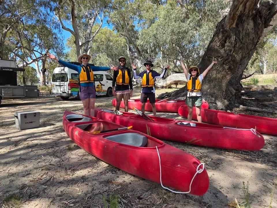 Four people standing on a sandy riverbank with red kayaks and paddles, wearing life jackets and hats, smiling and with arms raised. In the background are trees, a white van, and outdoor equipment.