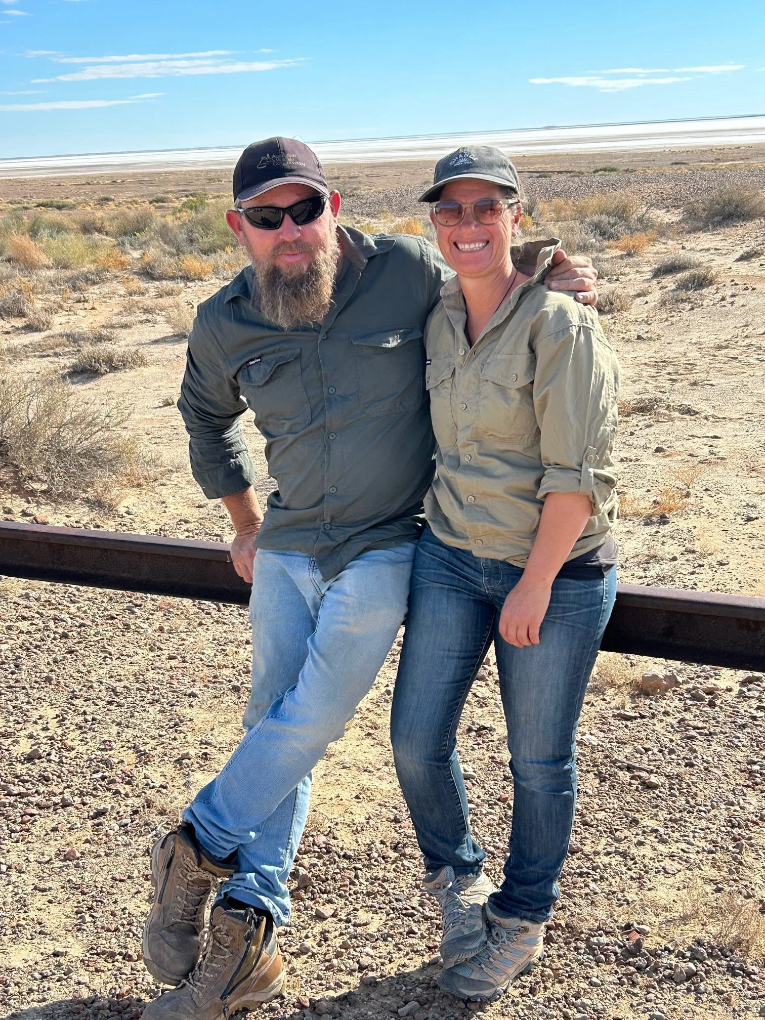 Tour guides Dan and Rian standing in a desert landscape, smiling at the camera, with a clear blue sky overhead.