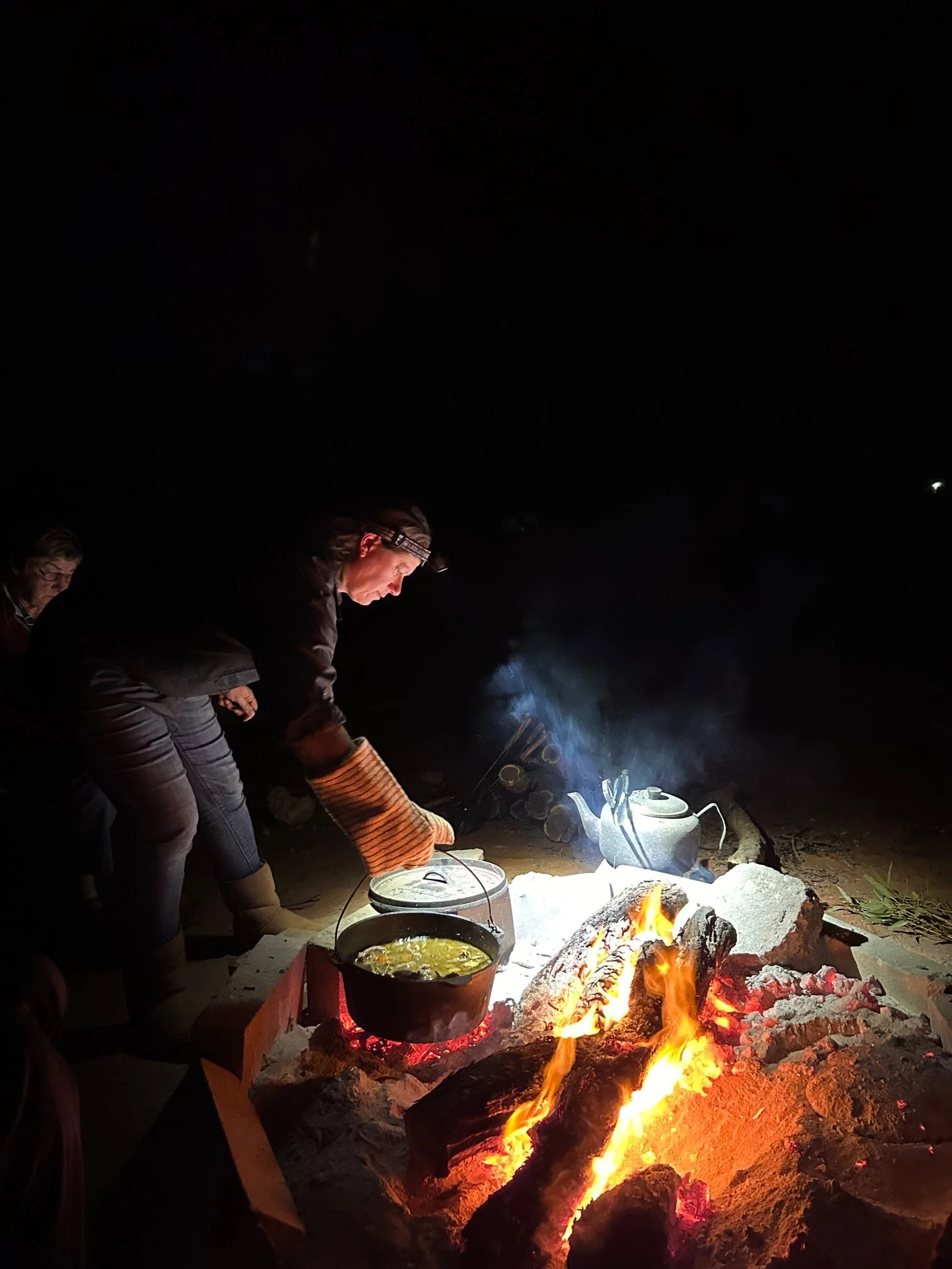 People cooking over an open campfire at night, with pots on the fire and logs burning.