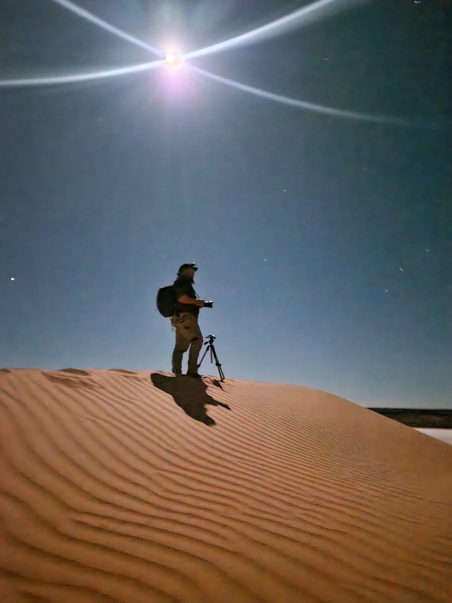 Person on sand dune at night with a camera and tripod, under bright moonlight with visible stars in the sky.