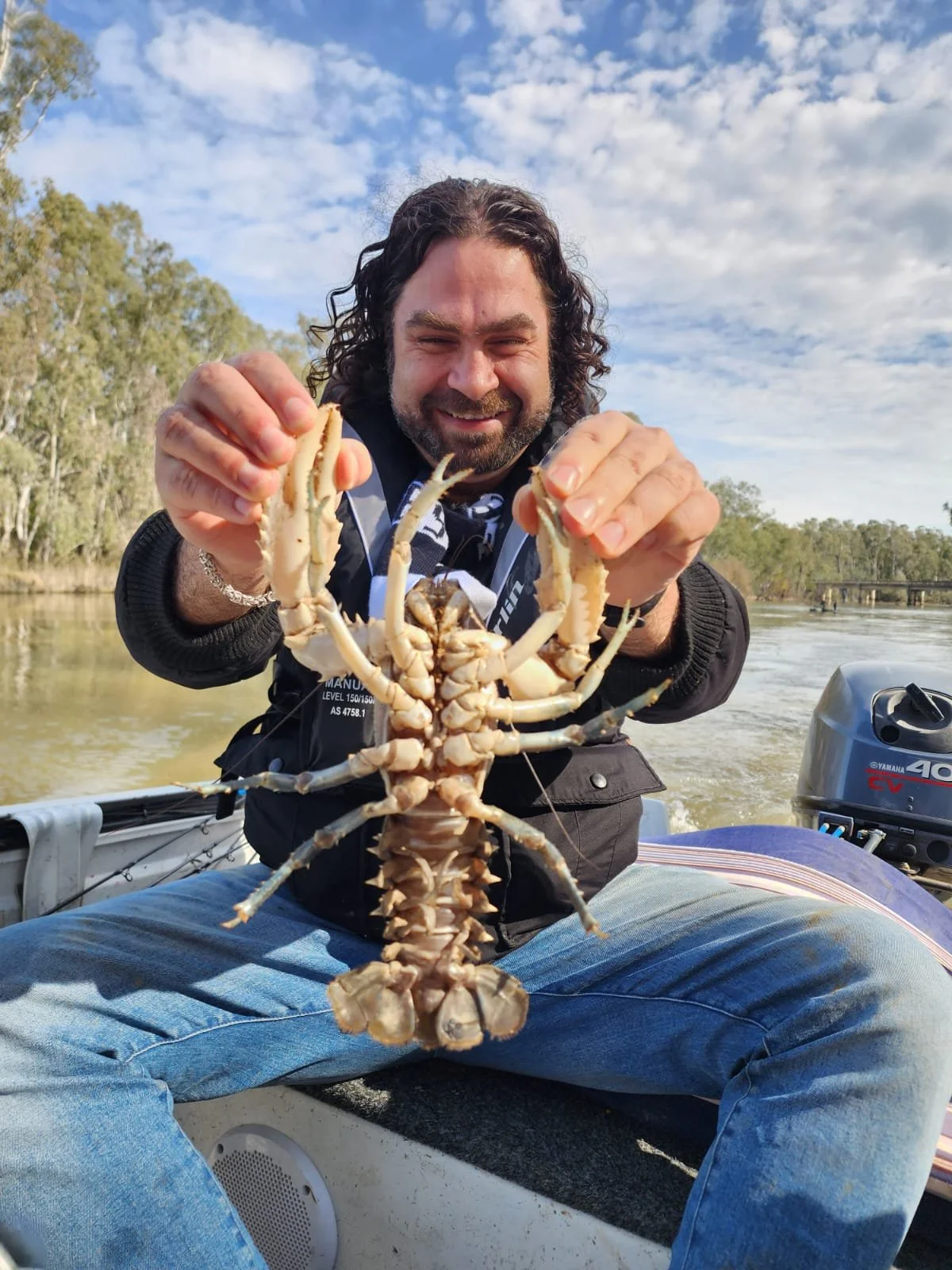 Man on a boat holding a large Yabby, smiling, with water and trees in the background.