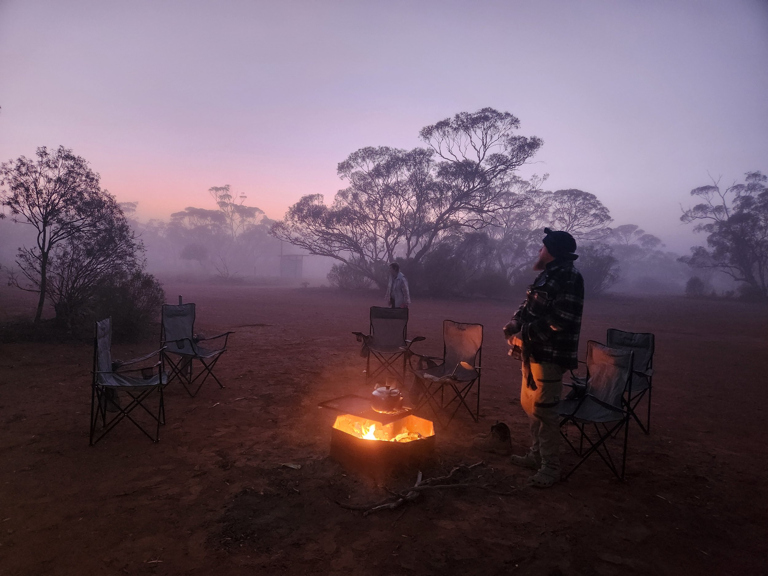 Two people at a campsite around a campfire in a foggy, wooded area at dawn or dusk, with trees in the background and empty chairs around the fire.