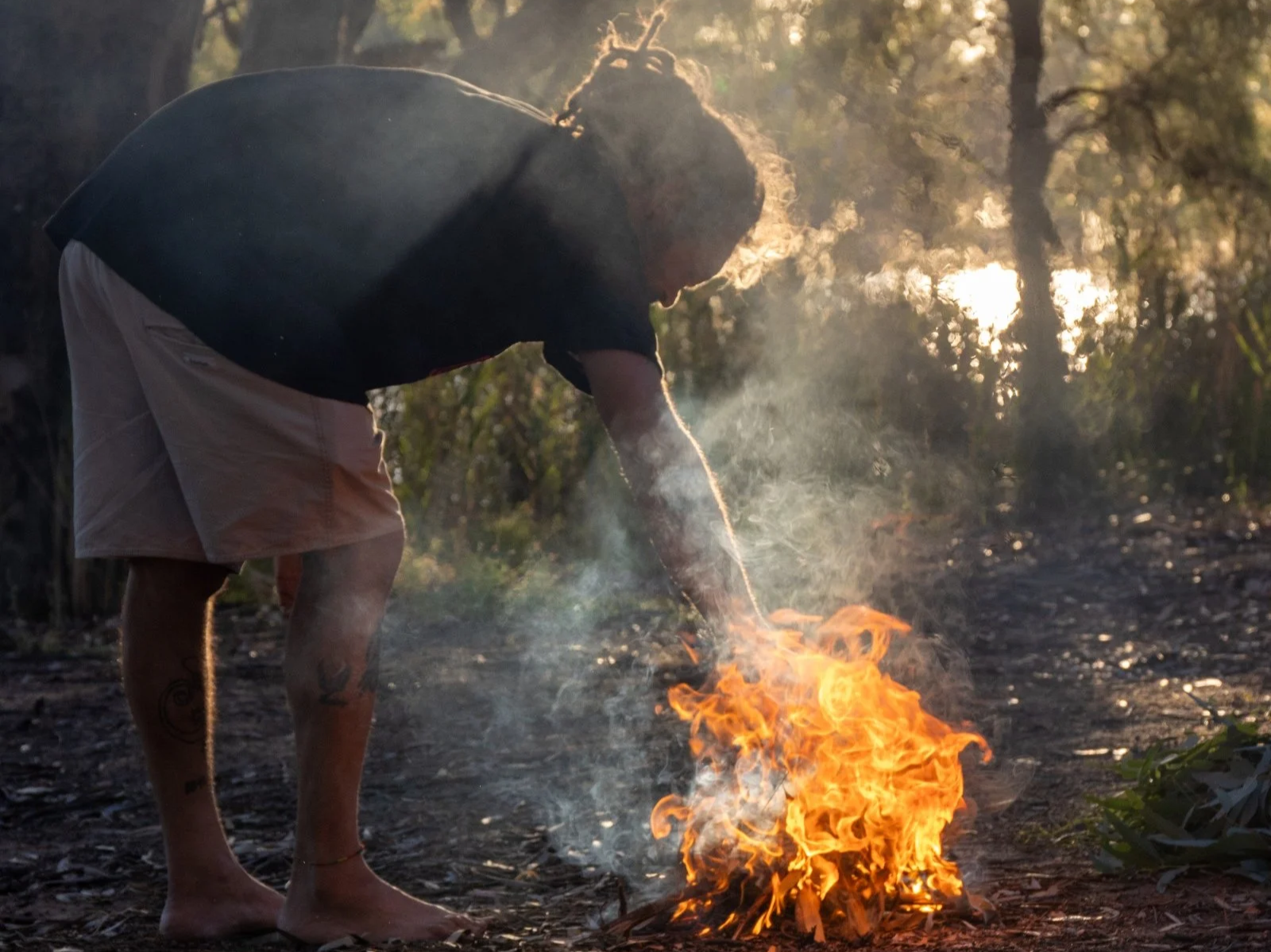 Person wearing a black t-shirt and beige shorts, with tattoos on their legs, standing outside during sunset, bending down near a small fire on the ground with flames visible and smoke rising, surrounded by trees and foliage.