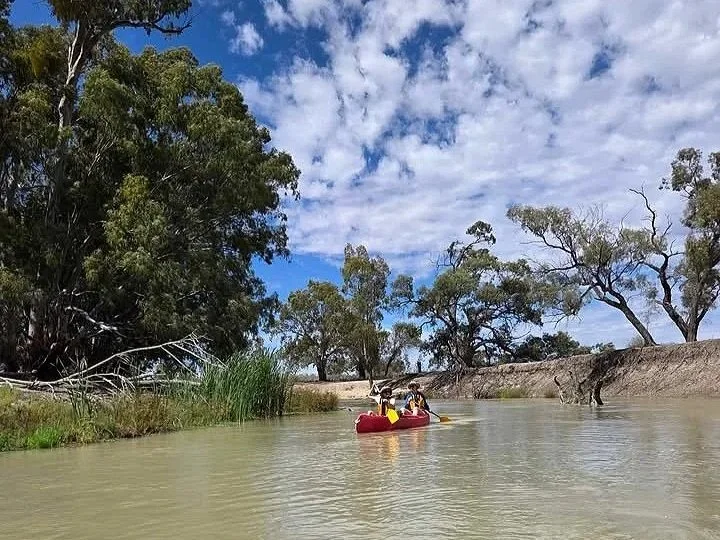 People kayaking on a river surrounded by trees with a partly cloudy sky.