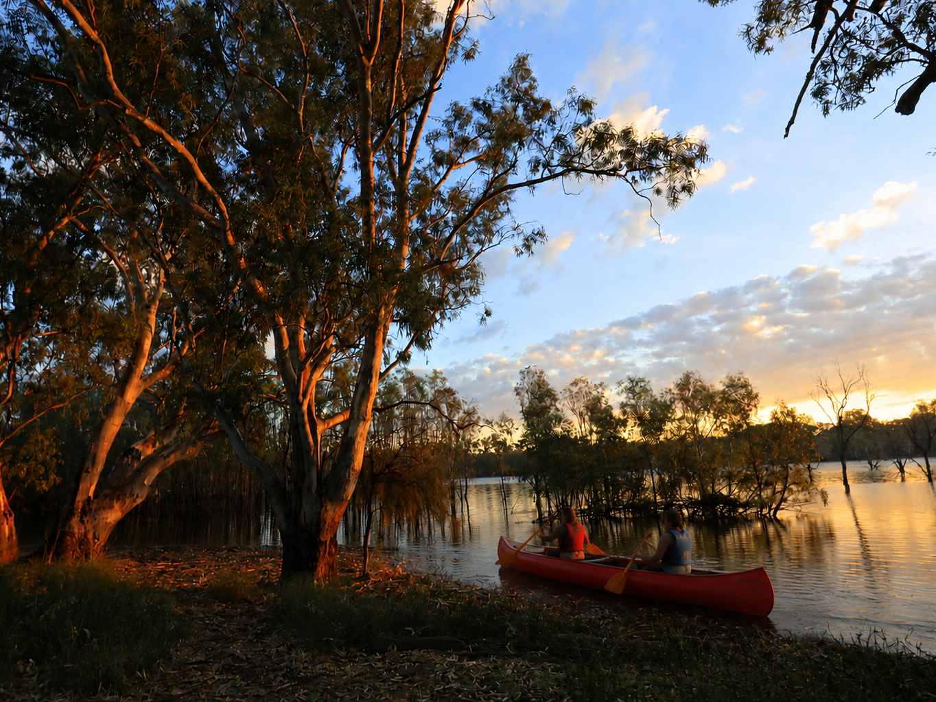 People kayaking on a calm river near trees during sunset, with a partly cloudy sky.
