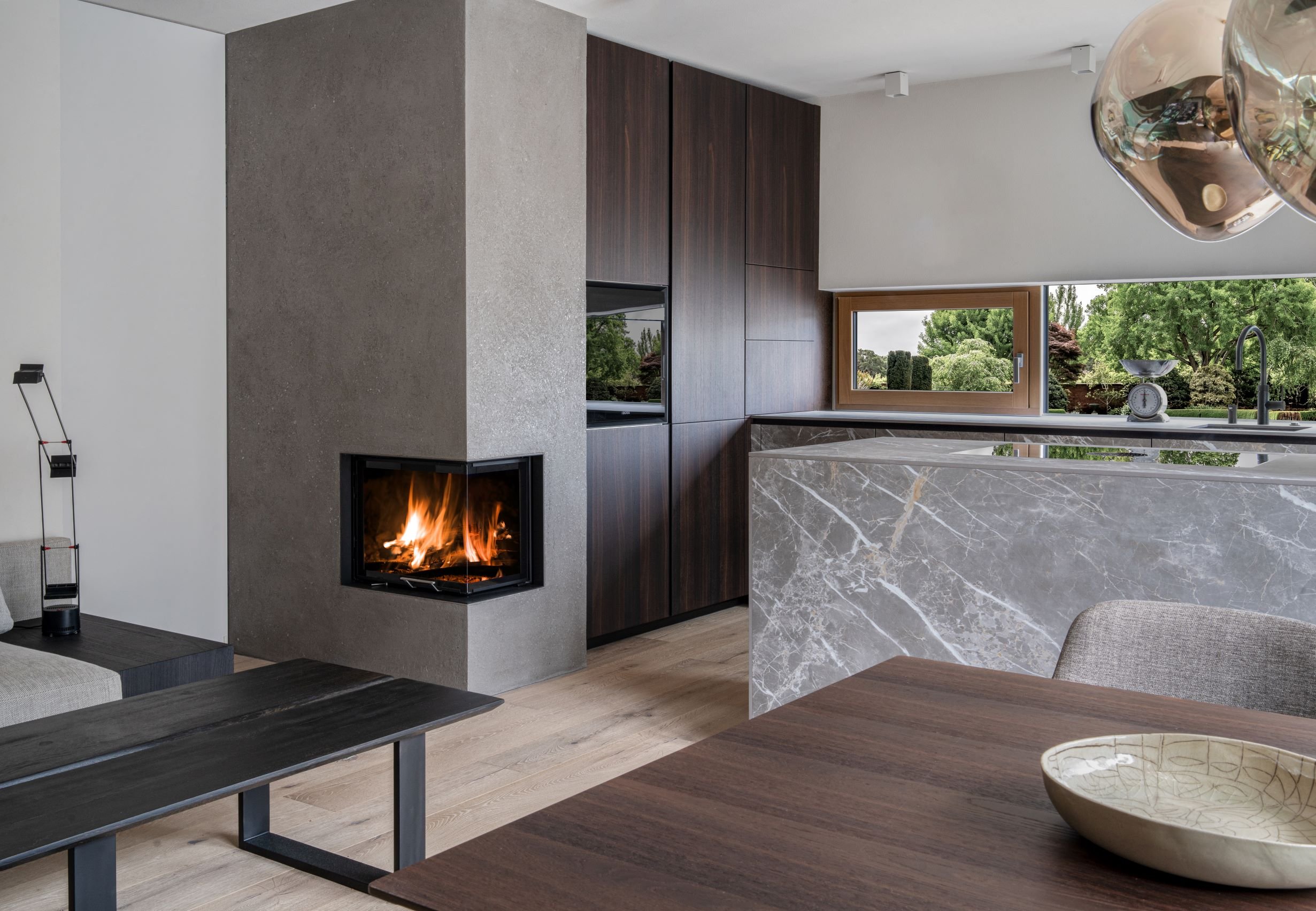 Innenansicht eines modernen Wohnzimmers mit Kamin, Holzdecken, großen Fenstern und Blick auf einen Garten mit Bäumen.