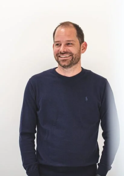Ein lächelnder Mann mit dunklem Pullover vor weißem Hintergrund.