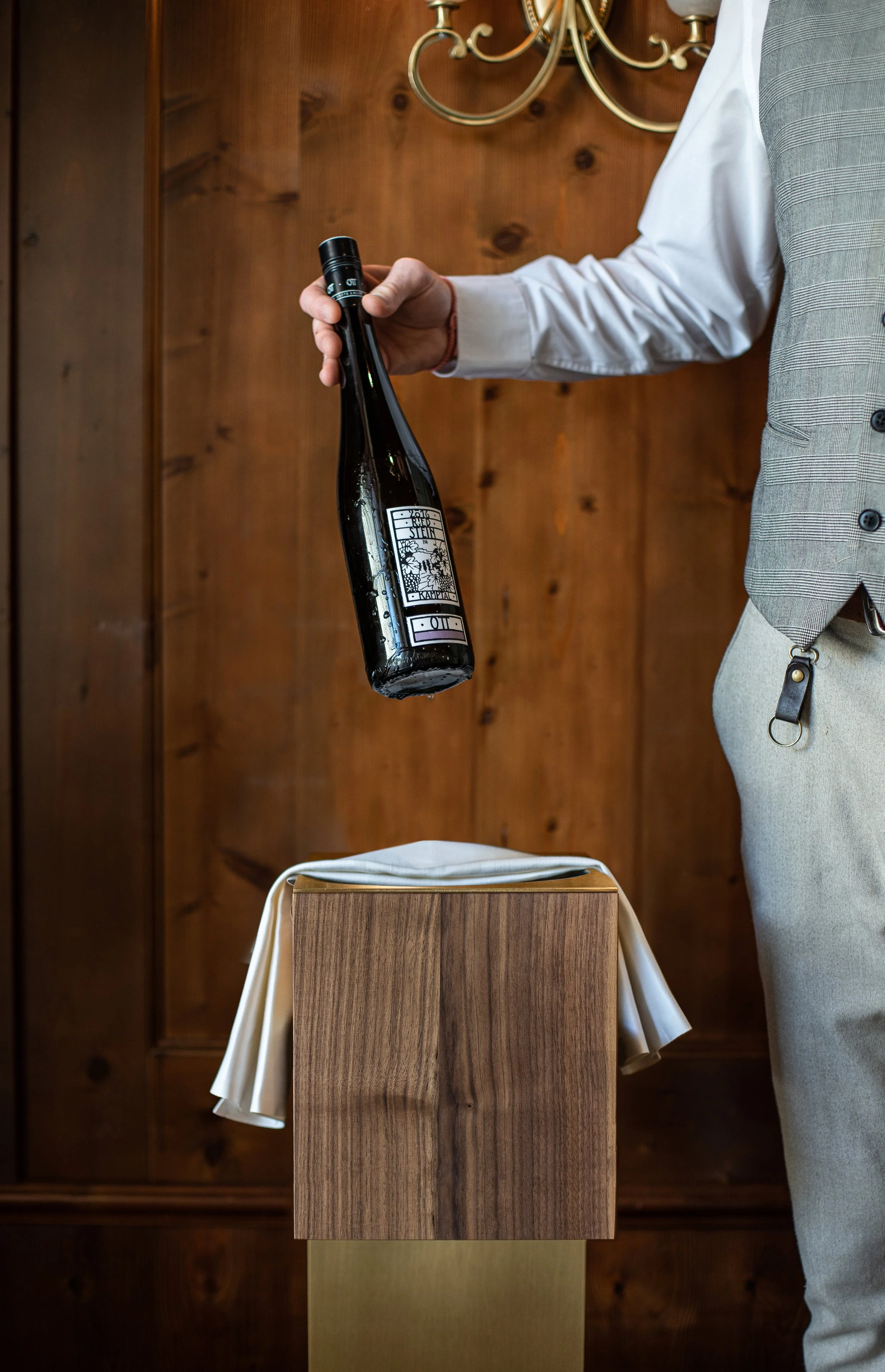 Man in Anzug und weißem Hemd hält eine Flasche Sekt über einem Holztisch, der mit einem weißen Tuch bedeckt ist, vor einer Holzvertäfelung und einer Deckenleuchte.
