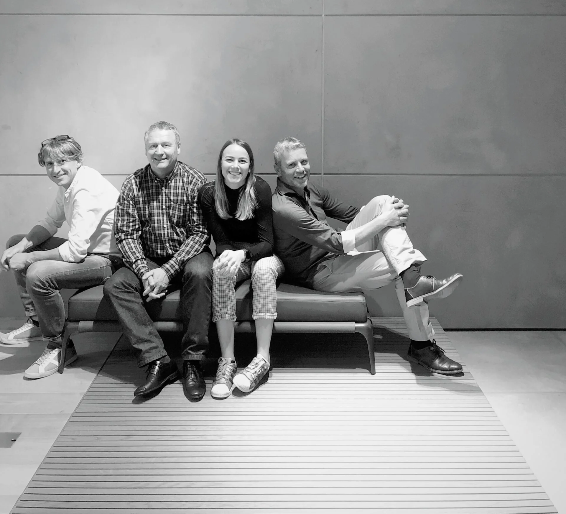 Fünf Personen sitzen auf einer Bank vor einer grauen Wand. Die Gruppe besteht aus drei Männern und einer Frau, alle lächeln und schauen in die Kamera. Sie befinden sich in einem modernen Innenraum.