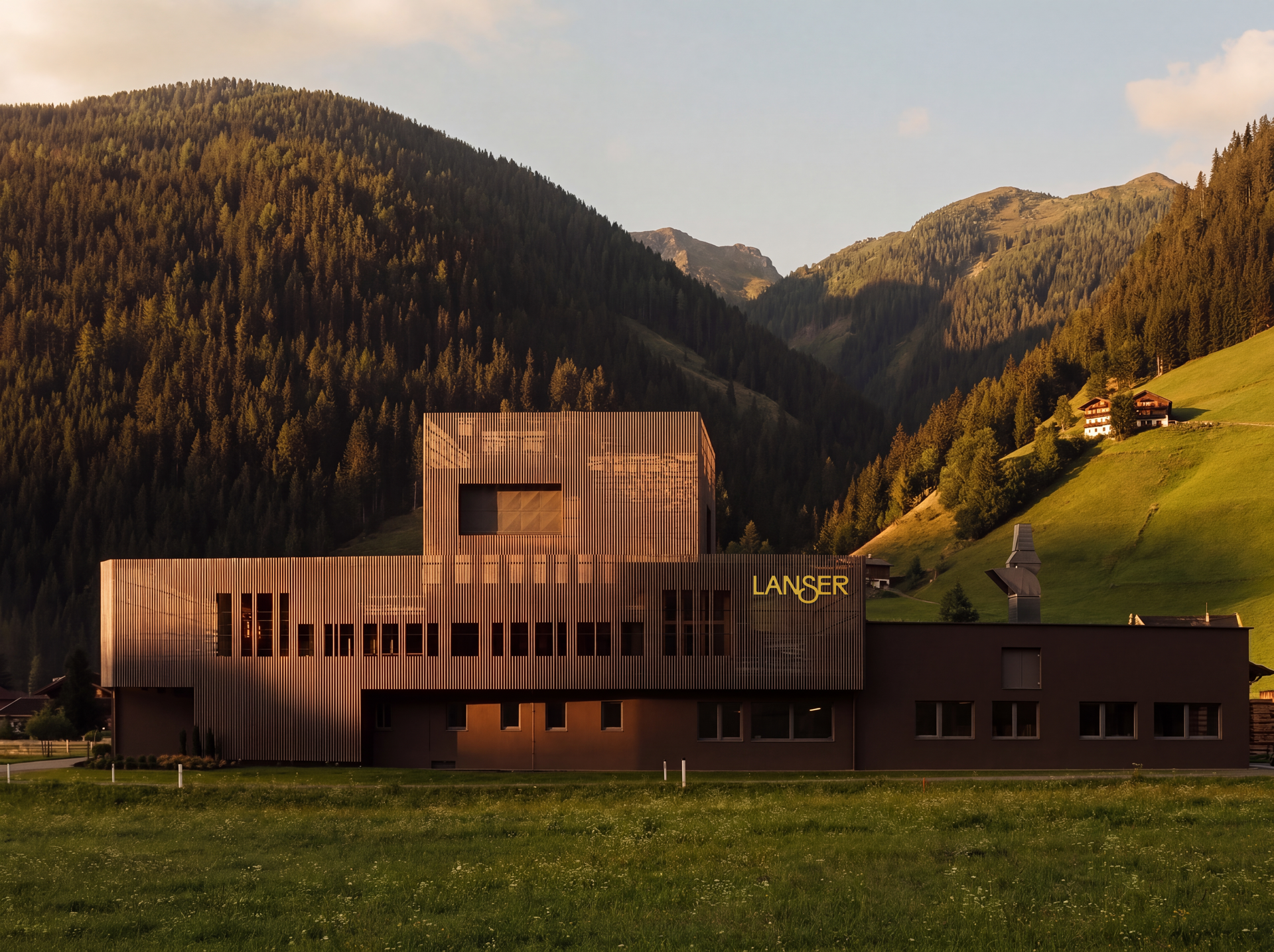 Modernes Gebäude mit Holzfassade im Vordergrund, umgeben von grüner Wiese, im Hintergrund Berge und Häuser, bei Sonnenuntergang.