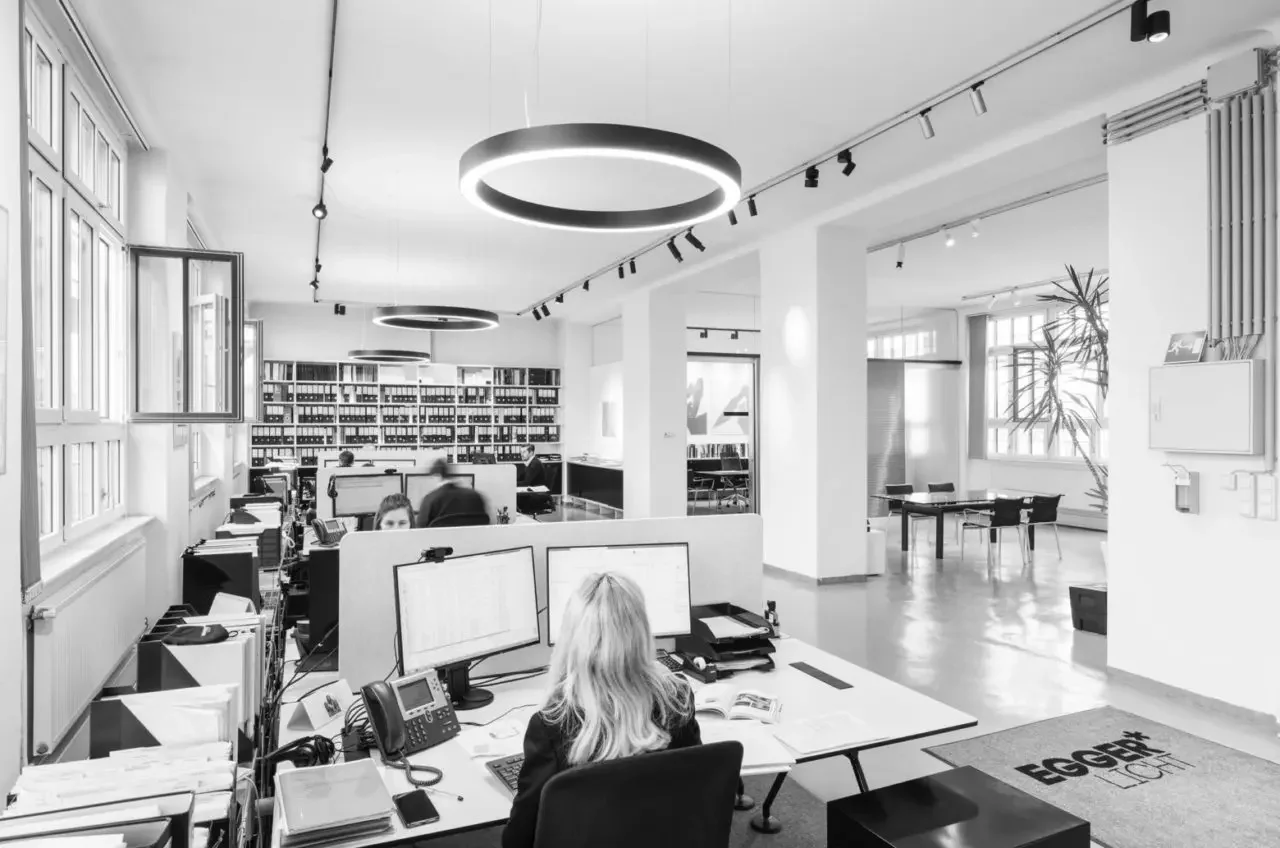 Moderne Büroarbeiterszene mit mehreren Mitarbeitern an Schreibtischen, große Fenster, Bücherregale im Hintergrund, Deckenleuchten und Pflanzen, helle Raumgestaltung.