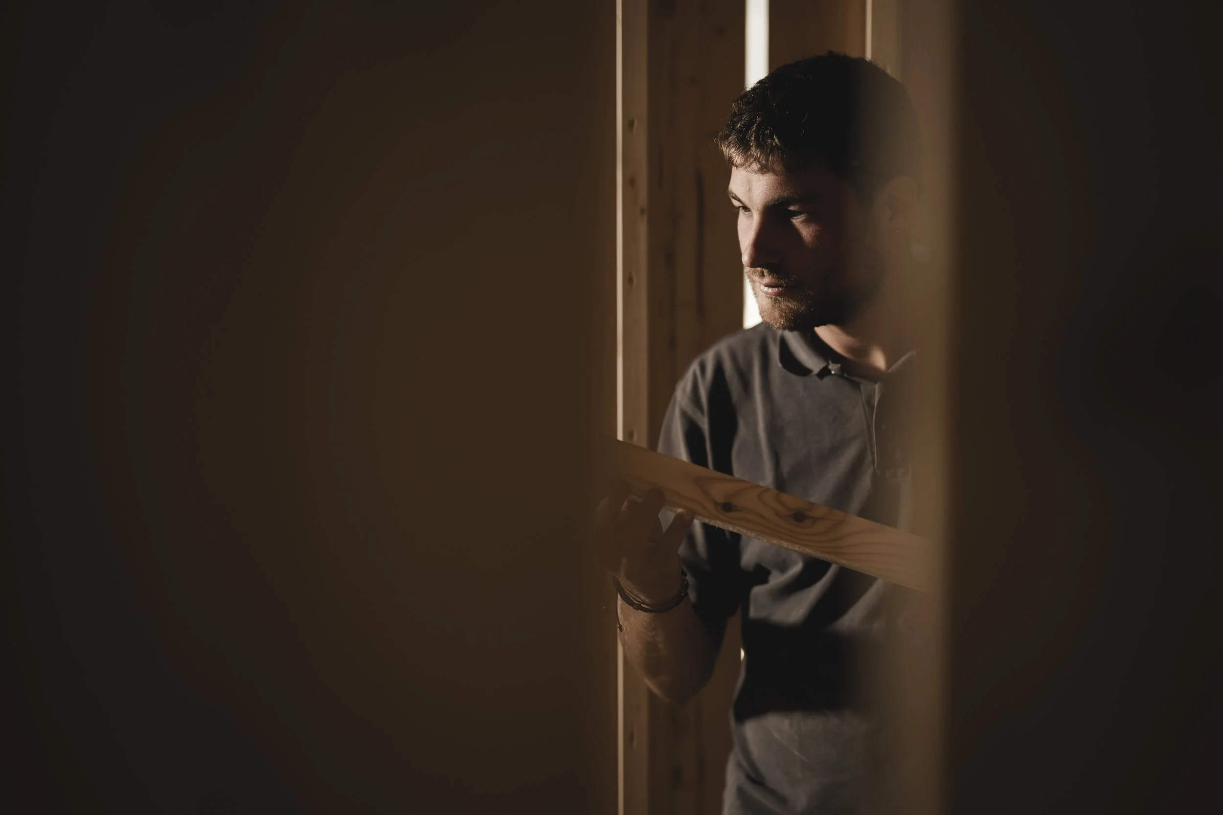 Mann betrachtet Holzstück beim Bau einer Wand, im Innenraum mit warmem Licht.