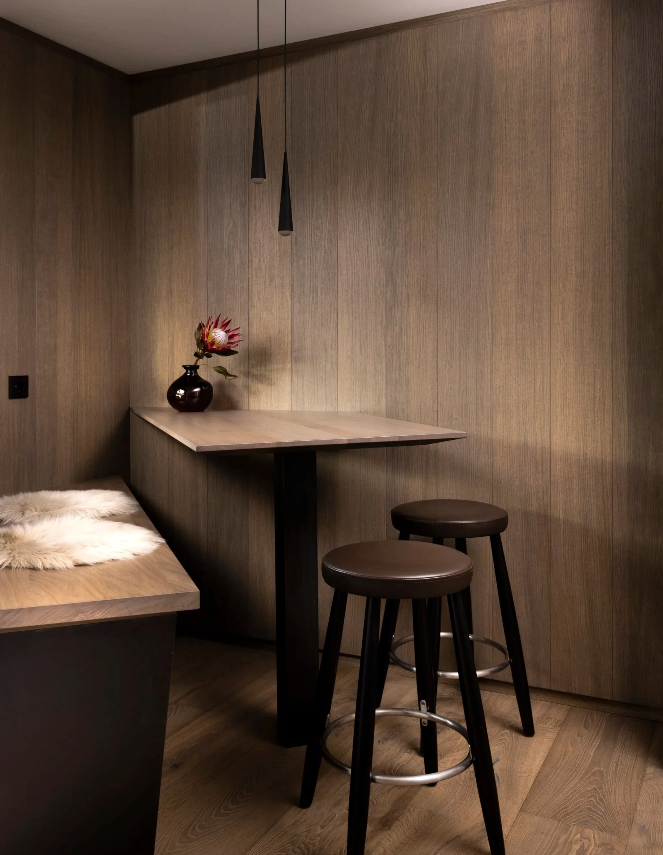 Ein minimalistischer Raum mit dunklen Hockern, einer Holzplatte und einer Vase mit einer Blume, Wandverkleidung aus Holz und zwei schwarzen Hängeleuchten.