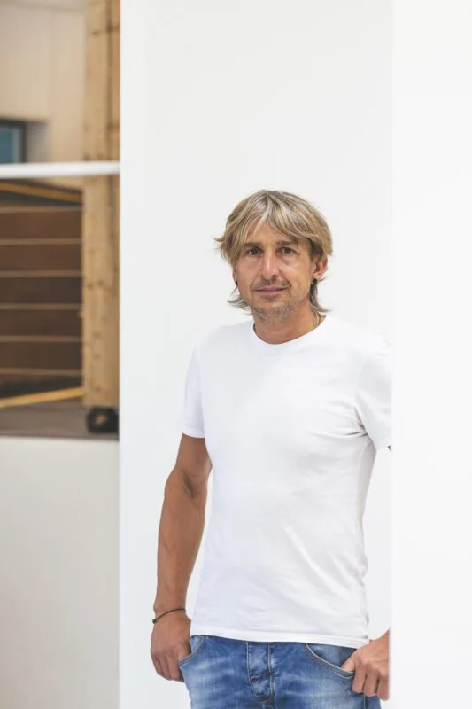 Ein Mann mit blondem, unordentlichem Haar, trägt ein weißes T-Shirt und blaue Jeans, steht vor einer weißen Wand in einem modernen Innenraum.