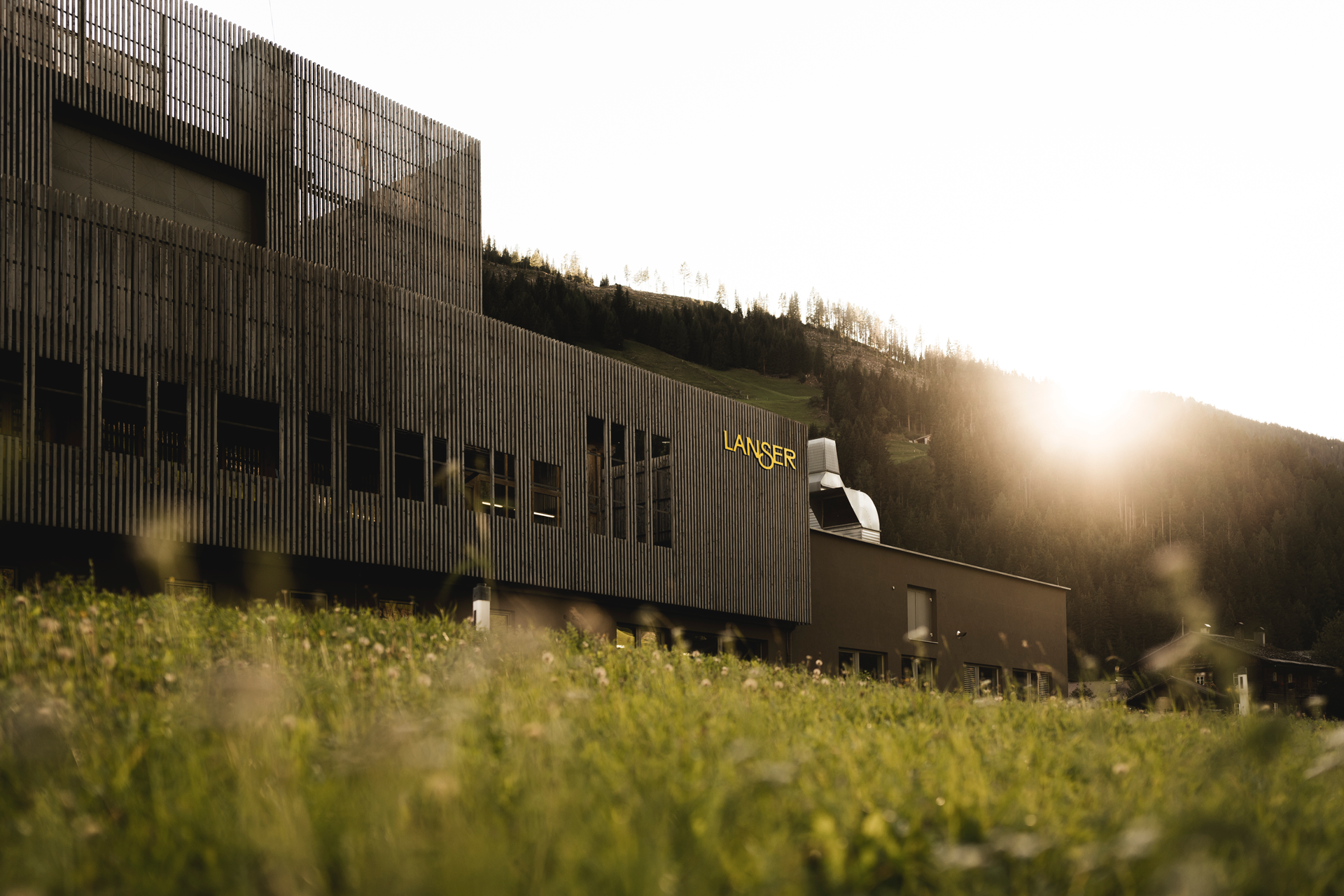 Moderne, dunkle Gebäudefassade mit vertikalen Holzlamellen, das Wort 'LANSER' in gelben Buchstaben an der Seite, umgeben von grünen Hügeln und Bäumen bei Sonnenuntergang.