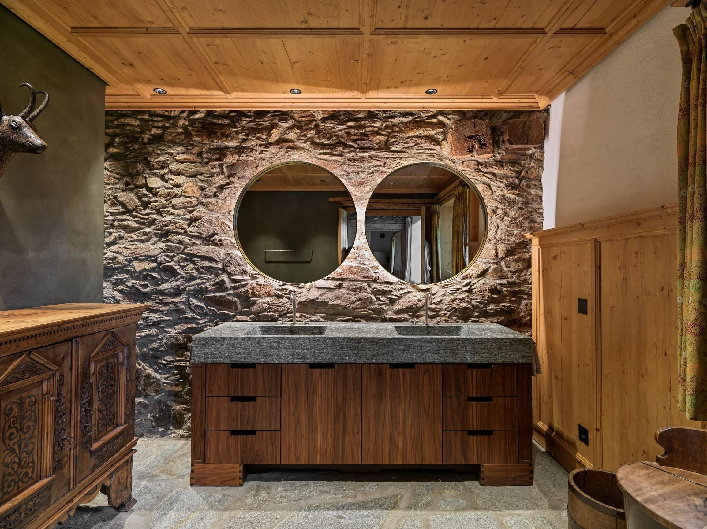 Ein rustikaler Raum mit einer Steinwand, einer Holzdecke und einem Doppelwaschbecken mit Spiegeln. Es gibt antike Holzmöbel und eine Gardine auf der rechten Seite.