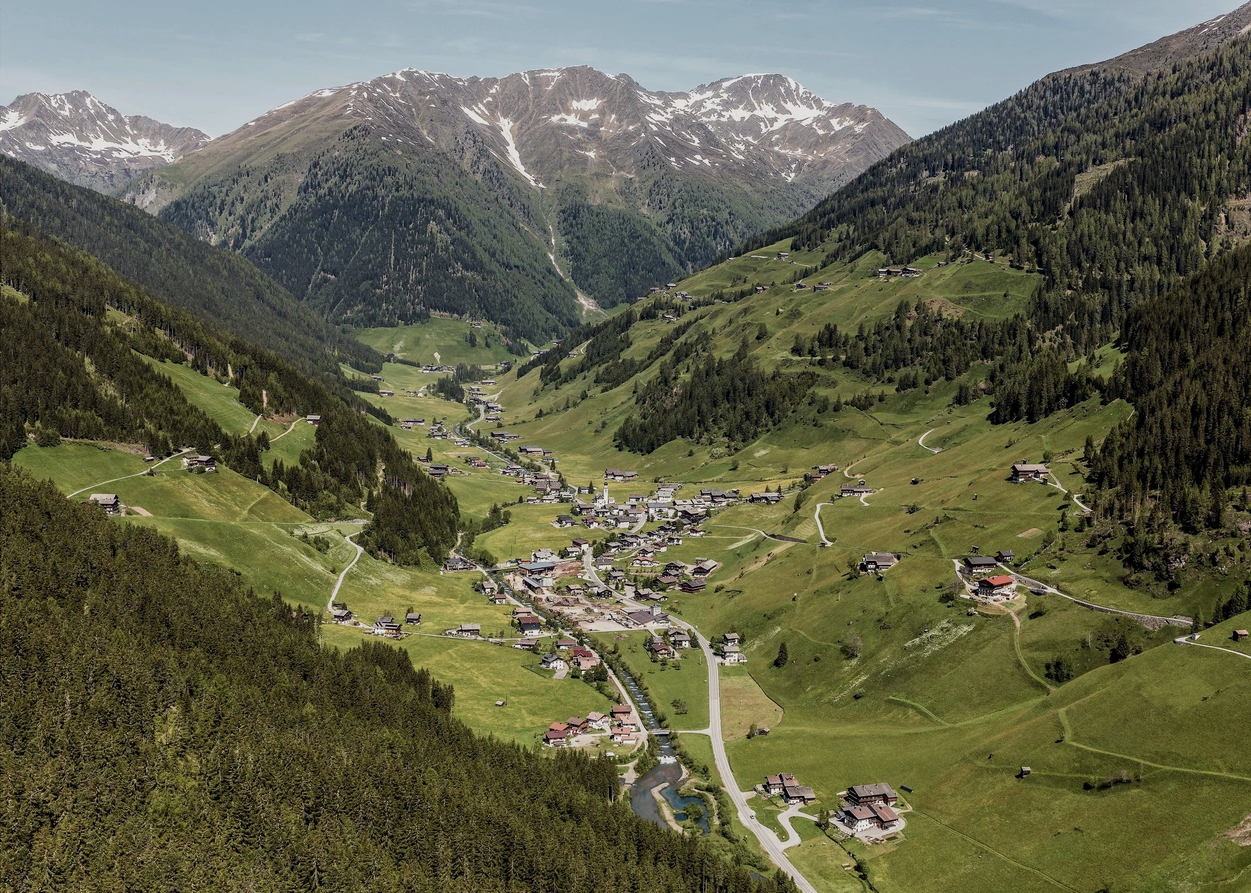 Alpenlandschaft mit grünen Hügeln, kleinen Dörfern, Wäldern und schneebedeckten Bergen im Hintergrund.