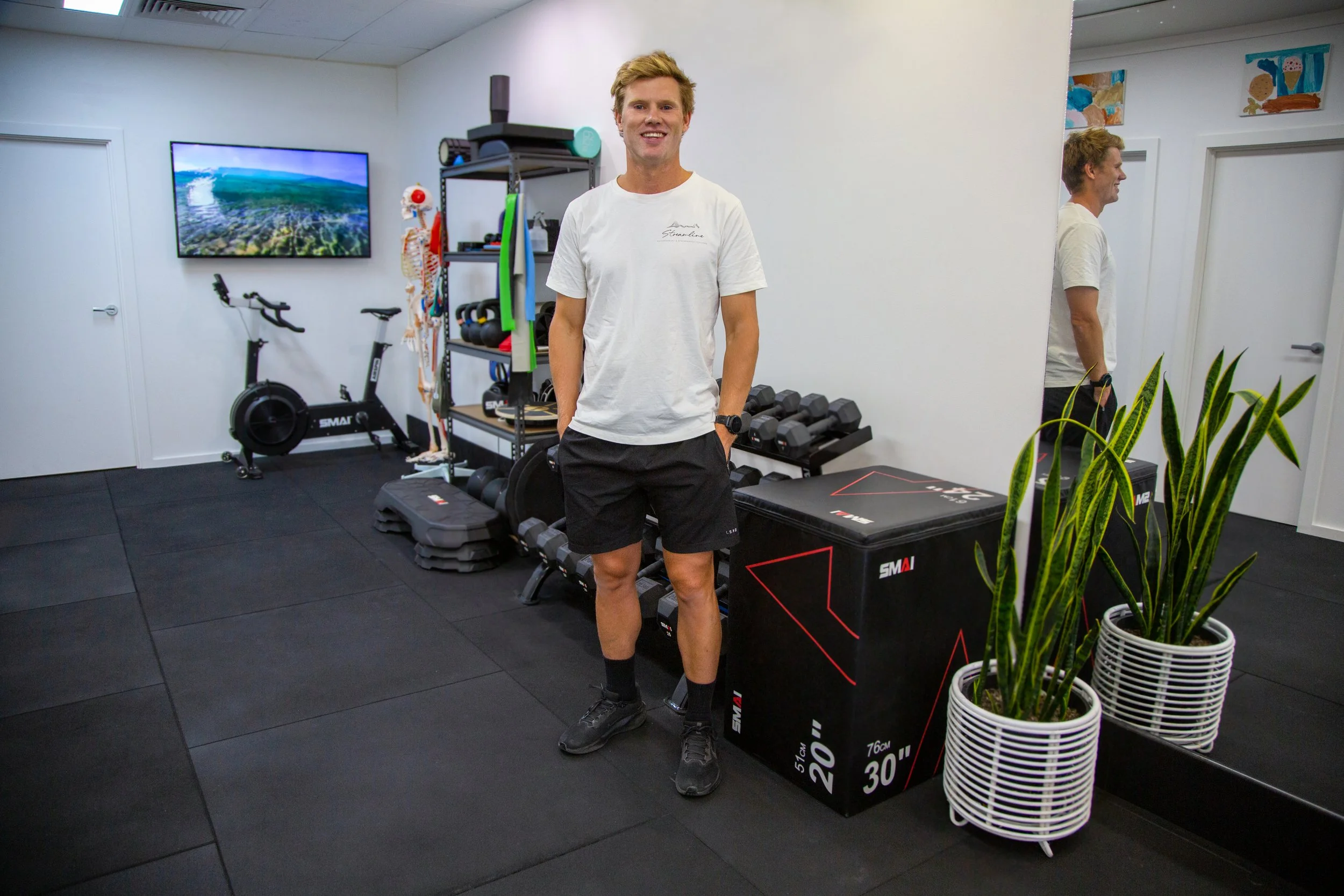 A young man with blonde hair, wearing a white T-shirt and black shorts, standing in a gym with fitness equipment, a large mirror, green plants, and colorful wall art.