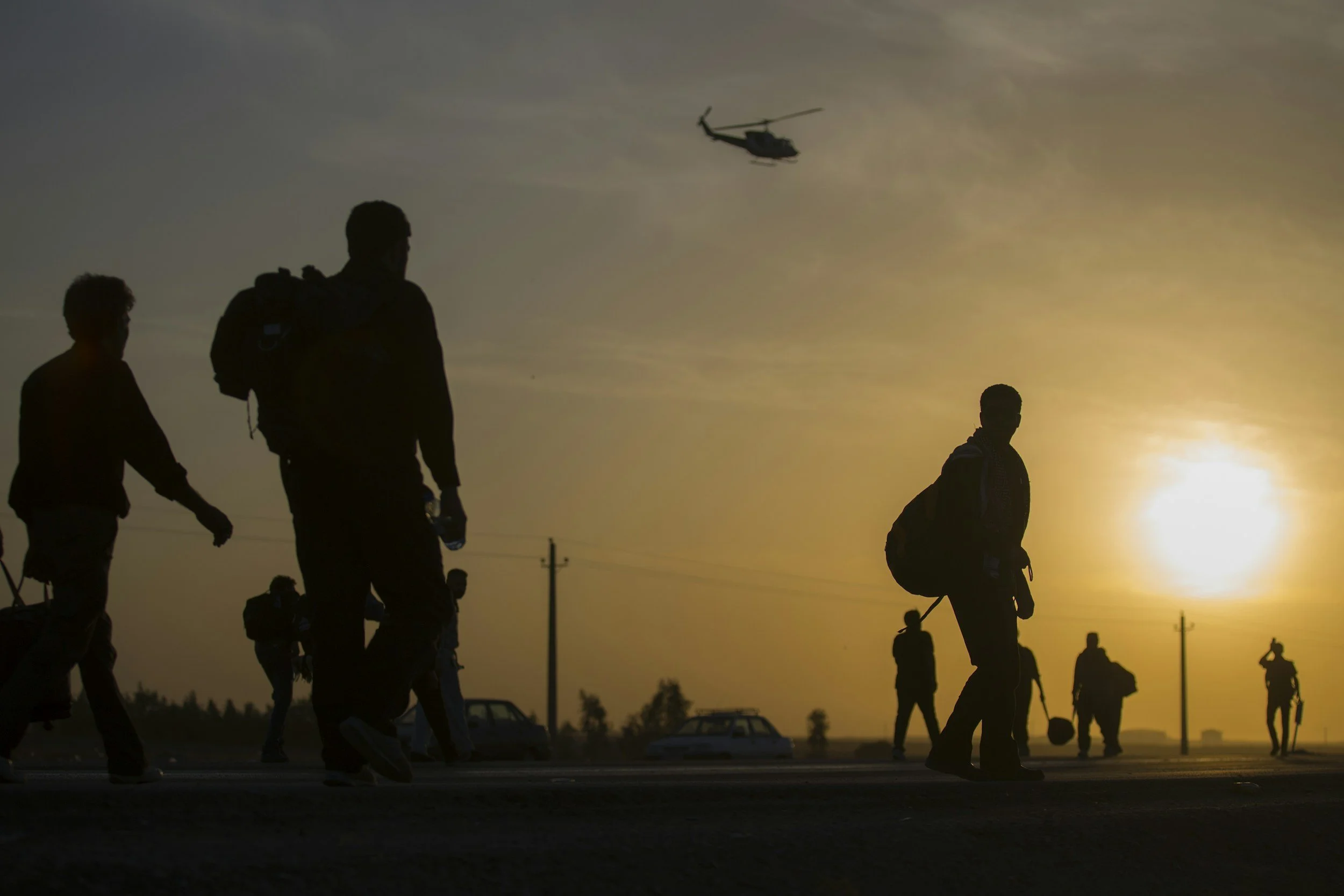 Silhouettes of people walking outdoors during sunset or sunrise with a helicopter flying in the sky.