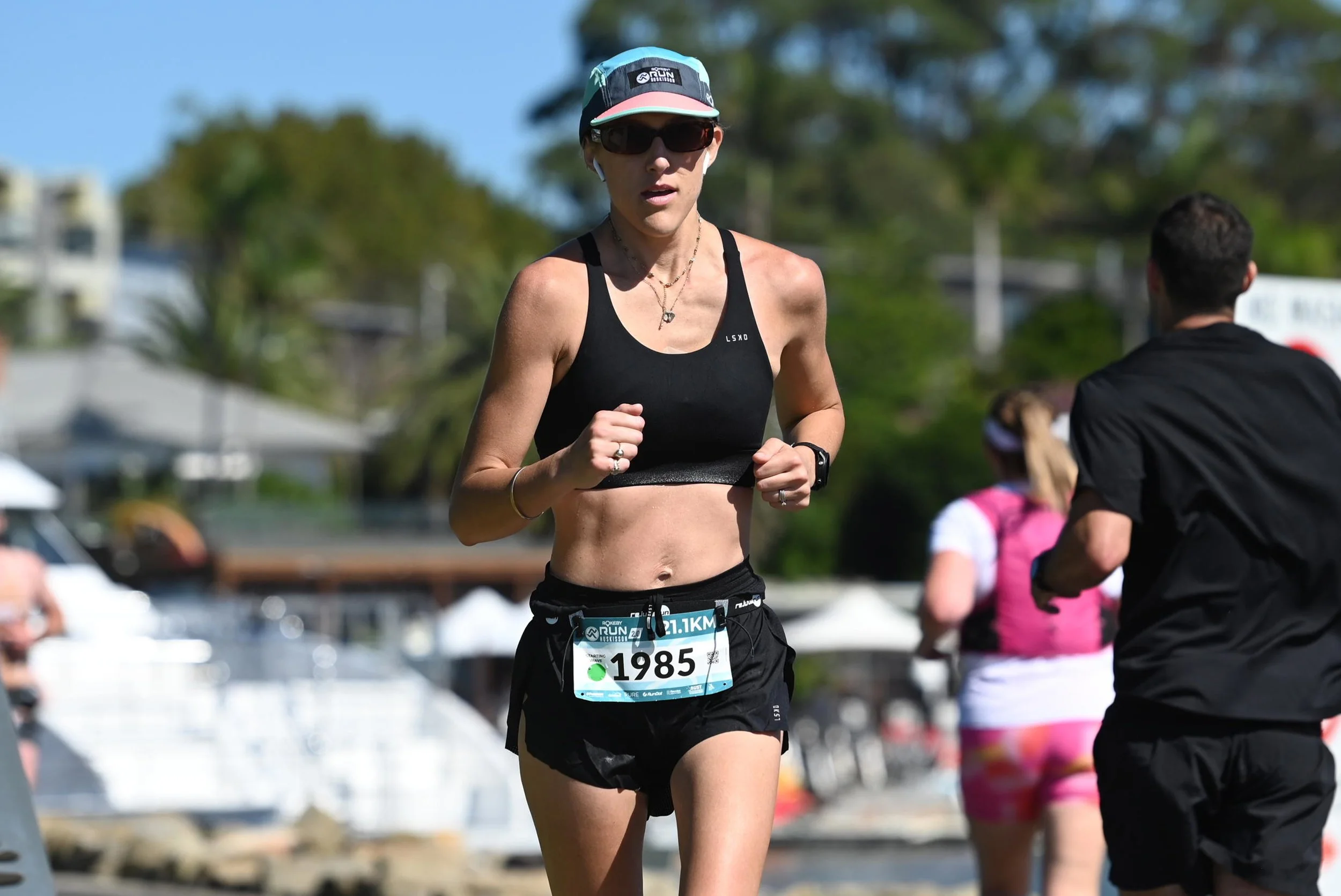 A female runner in black sportswear with sunglasses and a cap running in a race, with other runners and trees in background.