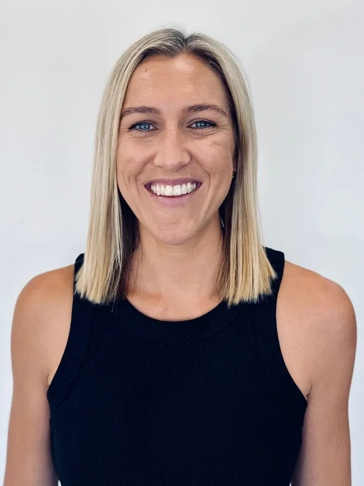 A woman with blonde hair, blue eyes, and a bright smile, wearing a black sleeveless top, standing against a plain white background.