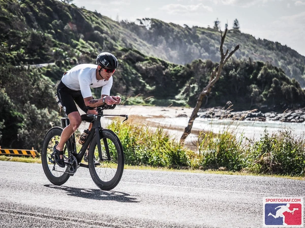A cyclist wearing a white shirt, black shorts, helmet, and sunglasses riding a black bike on a coastal road with greenery, a tree, and water in the background.
