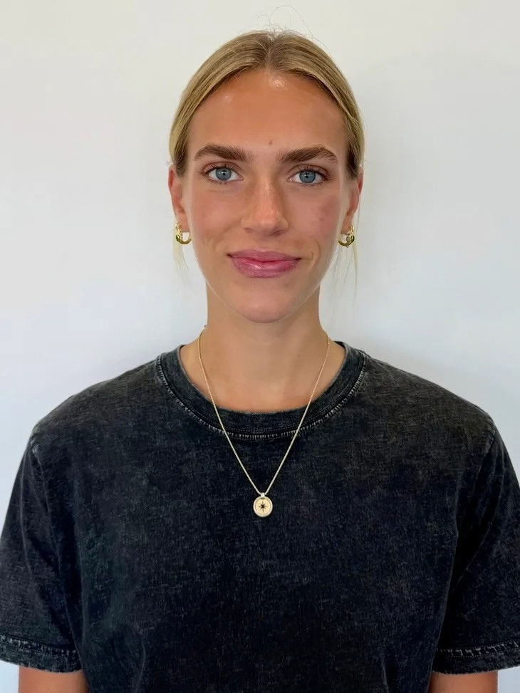 A young woman with blonde hair, blue eyes, and fair skin, wearing a black T-shirt, gold hoop earrings, and a necklace with a star charm, standing against a plain light-colored background.