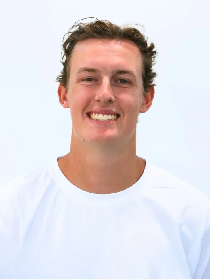 A young man with curly brown hair, fair skin, and a wide smile, wearing a white t-shirt against a plain, light background.