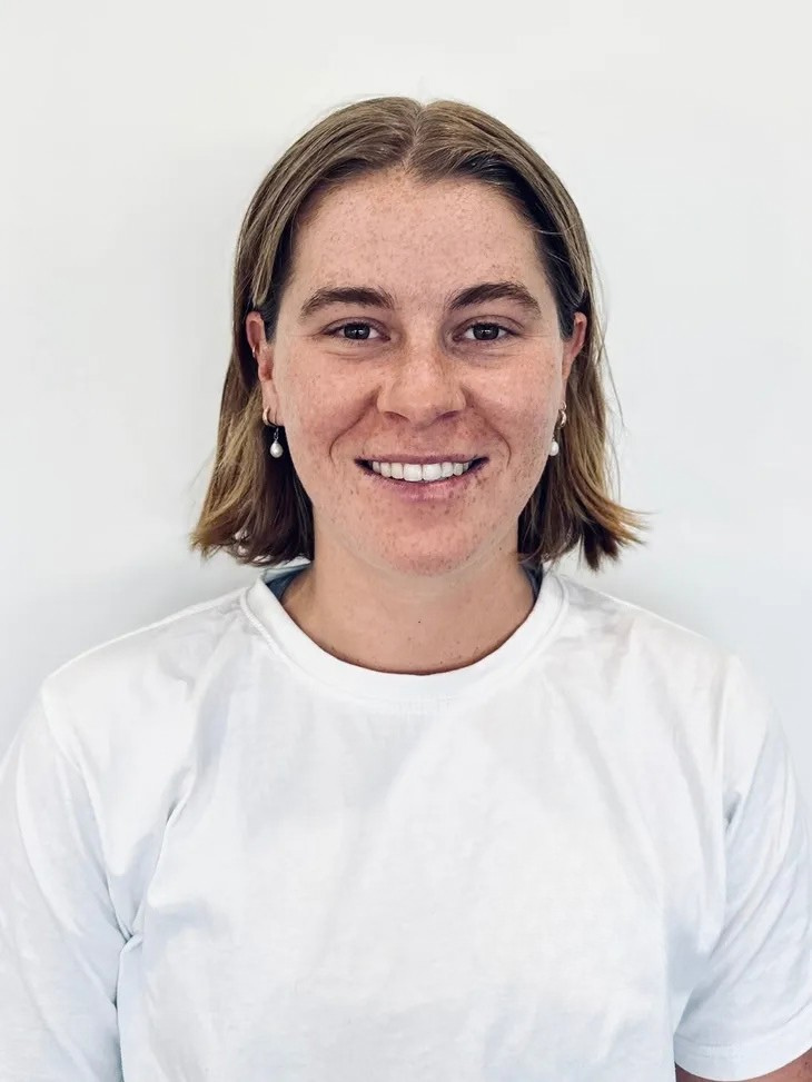 A young woman with shoulder-length light brown hair, freckles, and pearl earrings, smiling at the camera, wearing a white T-shirt against a plain white background.