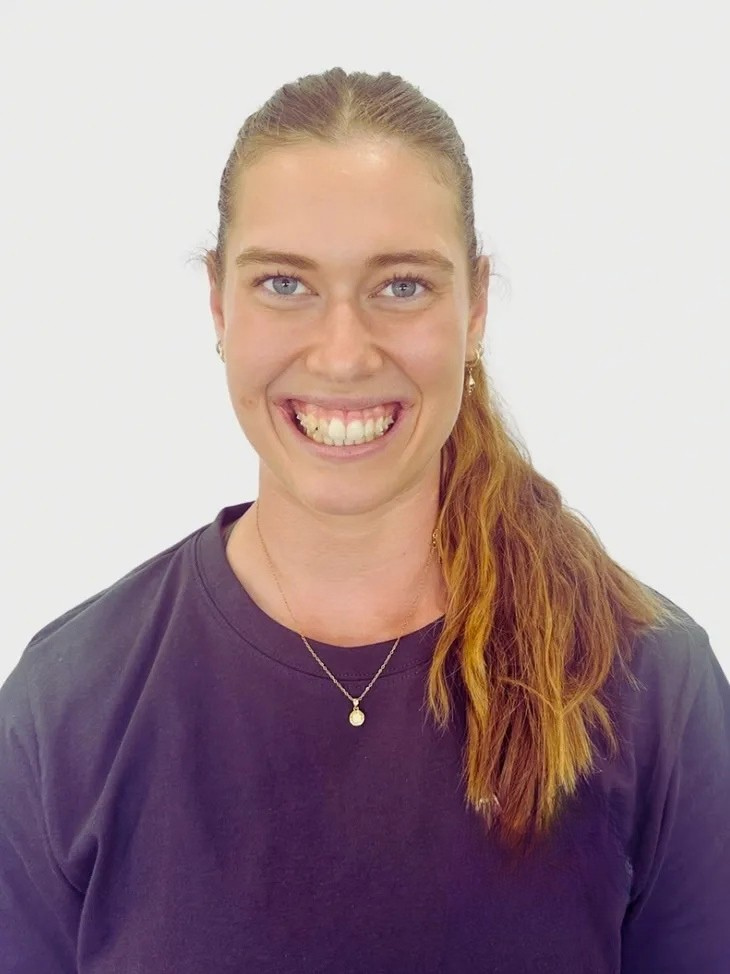 A young woman with blue eyes and reddish-brown hair in a ponytail, smiling at the camera, wearing a black shirt and a gold necklace with a pendant, against a plain white background.