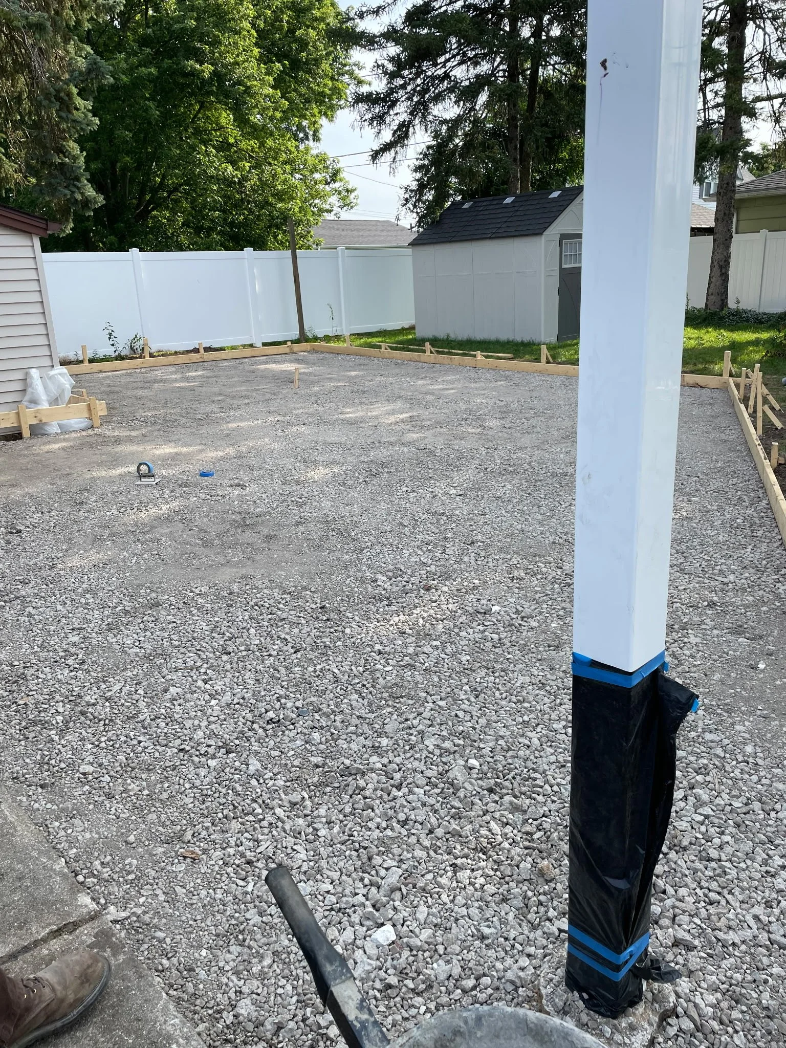 Construction site with a gravel ground, partially visible worker's boots, a white post with black and blue tape, and a white fence in the background with trees and a shed.