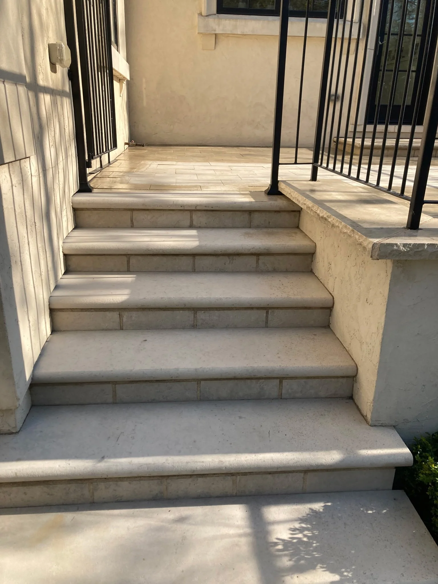 Exterior staircase with three white marble steps leading to a small landing, enclosed by black metal railings, with a beige stucco wall and a window in the background.