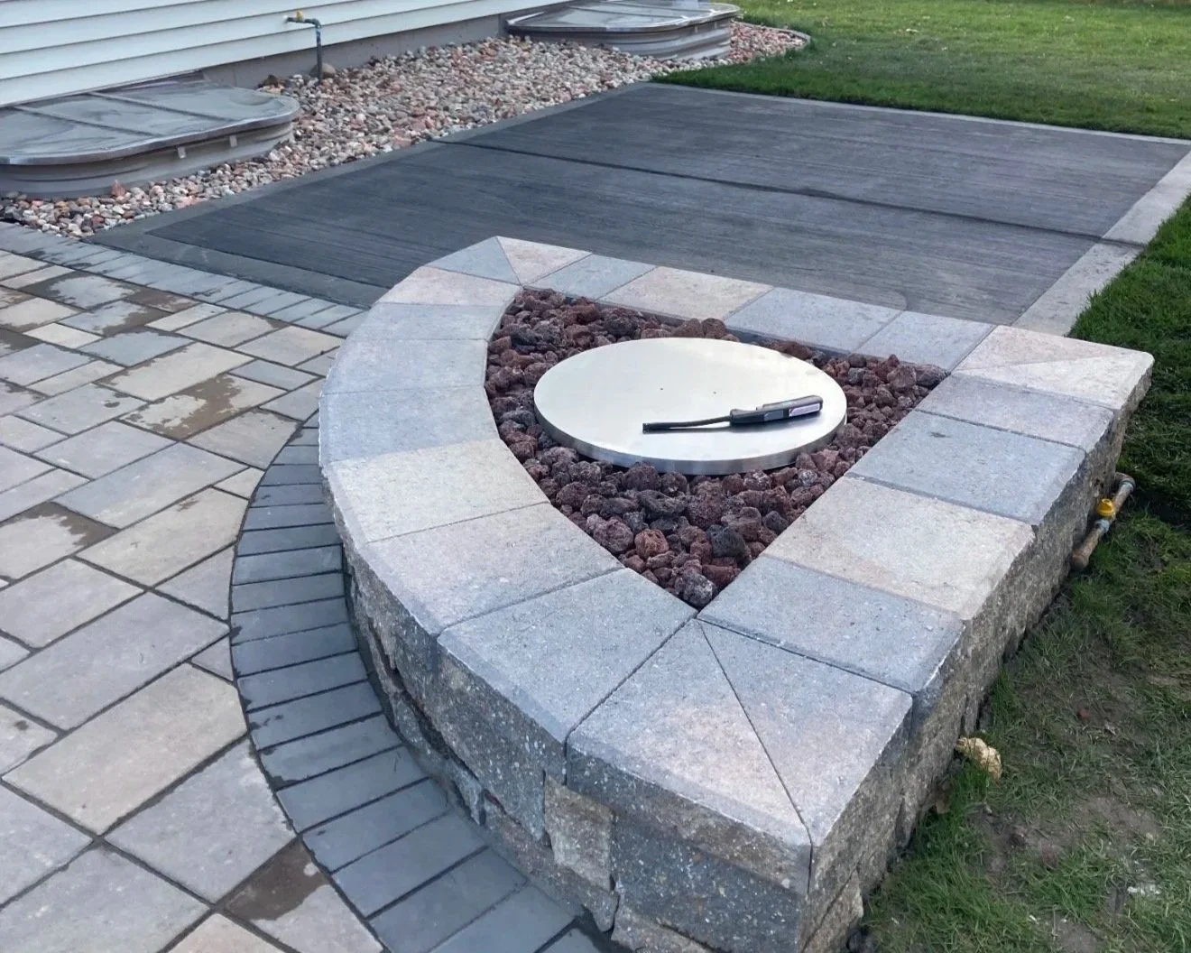 A stone fire pit with a round white insert and a gardening tool on top, surrounded by a low brick wall, with a patio made of large paver stones and a grassy yard in the background.