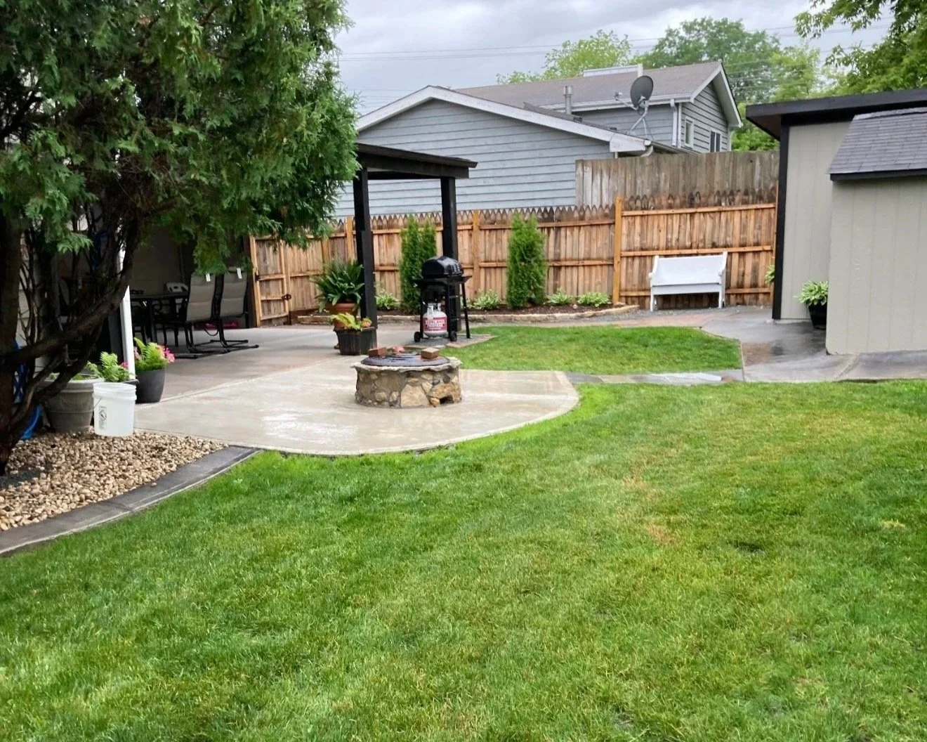 A backyard with a lush green lawn, a concrete patio area with a stone fire pit, a grill, potted plants, and a wooden fence enclosing the space. There are houses in the background.